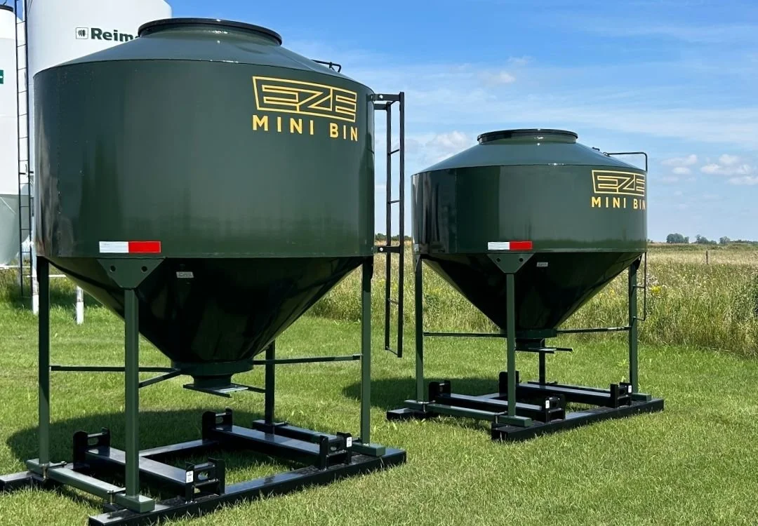 Two green mini-bin feed storage bins sitting on a grassy field with white Reimer Feed Bins in the backround.