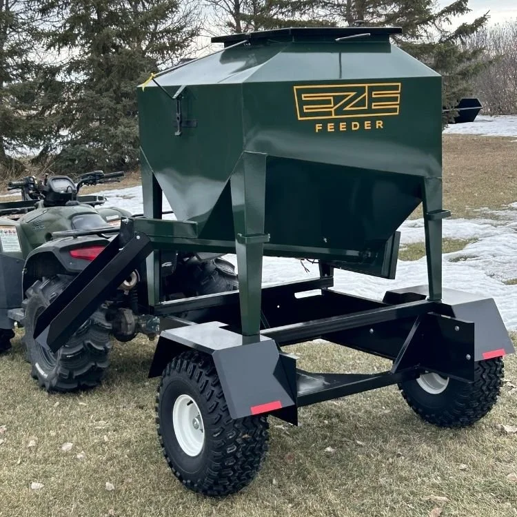 A dark green EZE Feeder with wheels hooked up to an ATV, used for dispensing feed for livestock, outdoors on a grassy area with some snow, trees, and cloudy sky in the background.