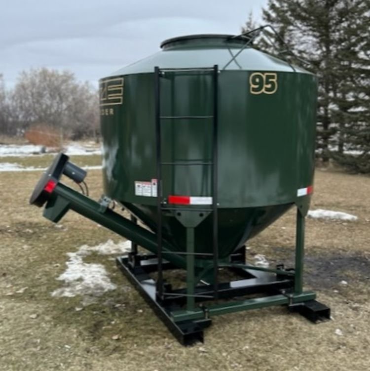 A green livestock feeder sitting in field with patches of snow on the ground and evergreen trees in the background