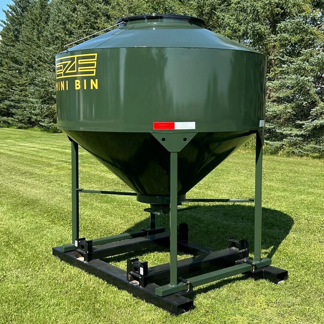 Side view of a green EZE Mini Bin farm storage tank sitting on green grass with evergreen trees in a row behind it.