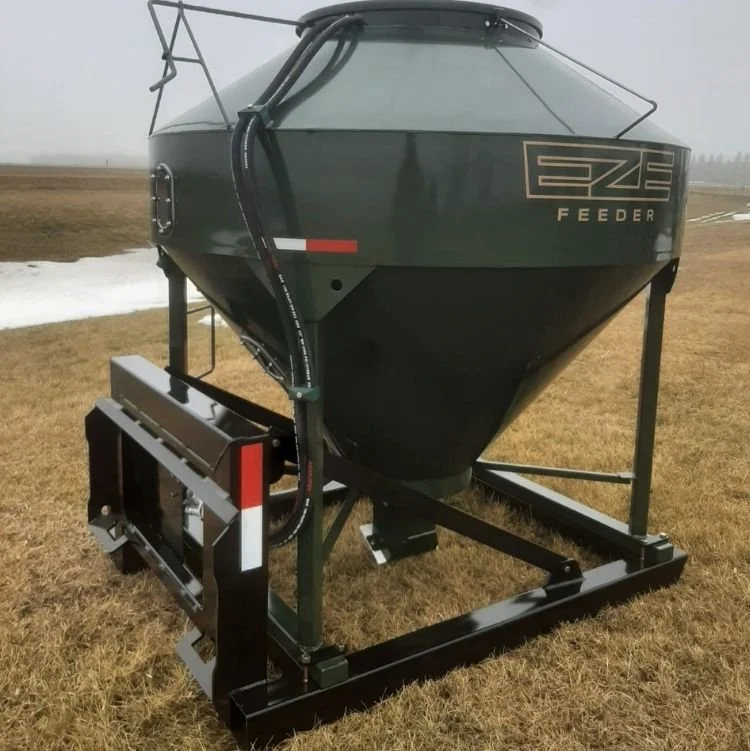 A green feeder with skid steer frame sitting on a field with dry grass, patches of snow and a foggy sky
