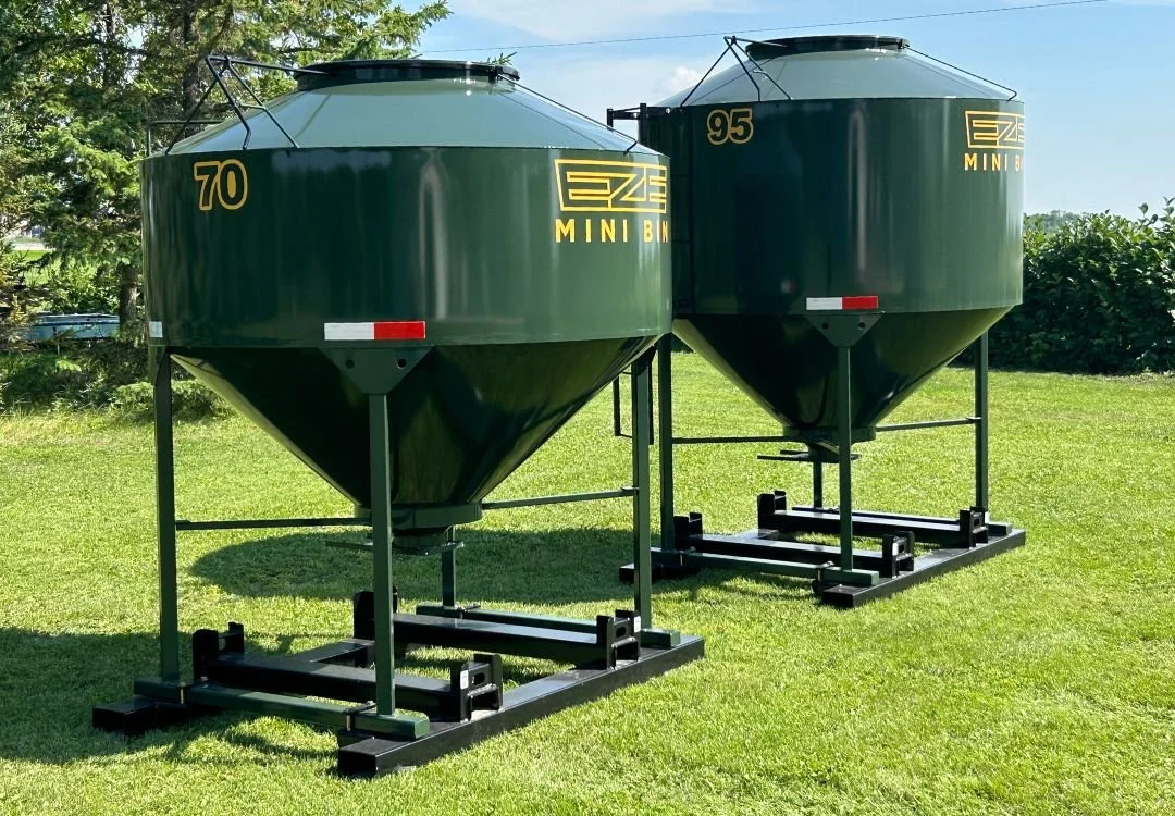 Two Eze-Feeder green steel feed storage bins, standing side by side on green grass with trees in the background.