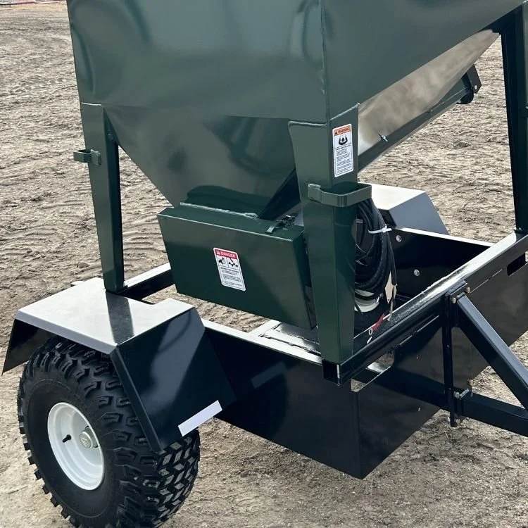 Close-up of a green metal industrial hopper on a black trailer with rugged tires, placed on dirt ground.