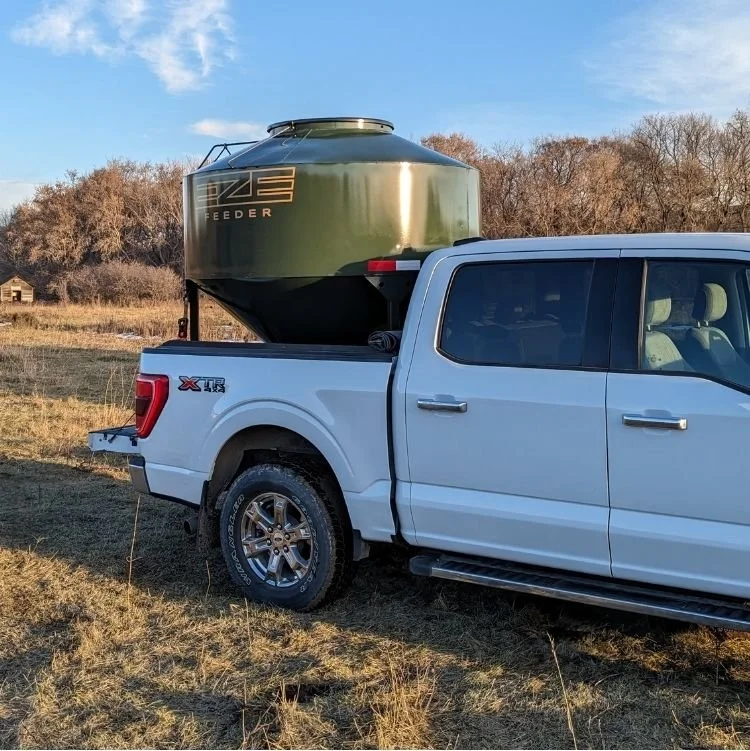 A white ford pickup with a green Eze-Feeder 70 bushel grain feeder in the truck box, sitting in a field with dry grass and bare trees in the background