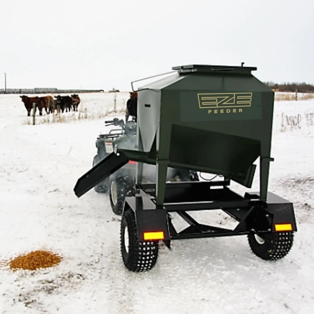 A small cattle feeder towed by a quad in a snowy field with cows in the background