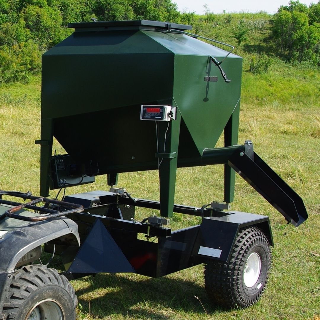 A  side view of a UTV 15 feeder on a trailer hooked up to a grey quad, parked in a grassy field with trees in the background
