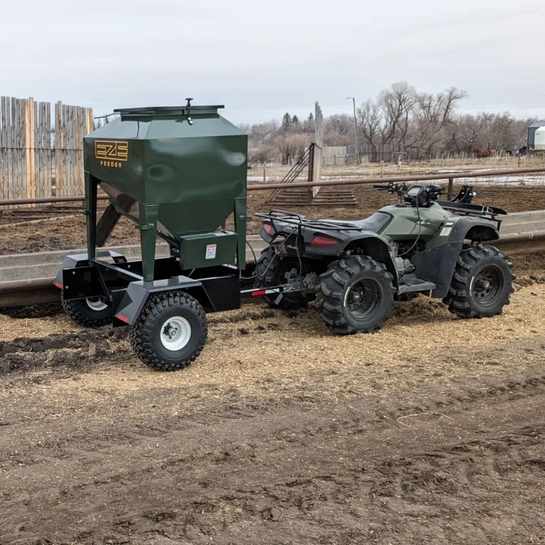 A  side view of a UTV 15 feeder on a trailer hooked up to a grey quad, parked in a muddy field with bare trees in the background