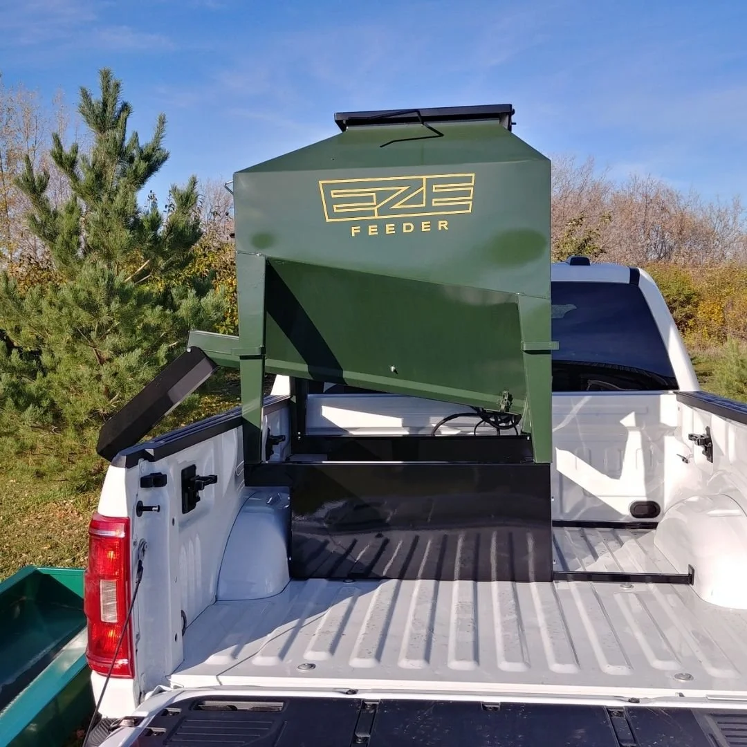A small green eze-feeder UTV cattle feeder loaded in the back of a white truck bed.