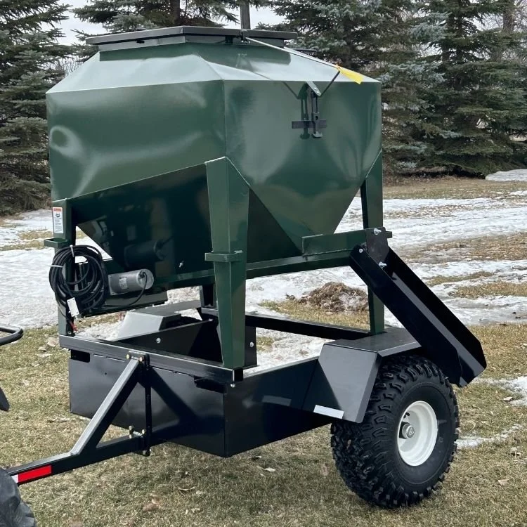 A small portable green ATV livestock Feeder with wheels sitting on a grassy field with snow in the background and trees in the background.