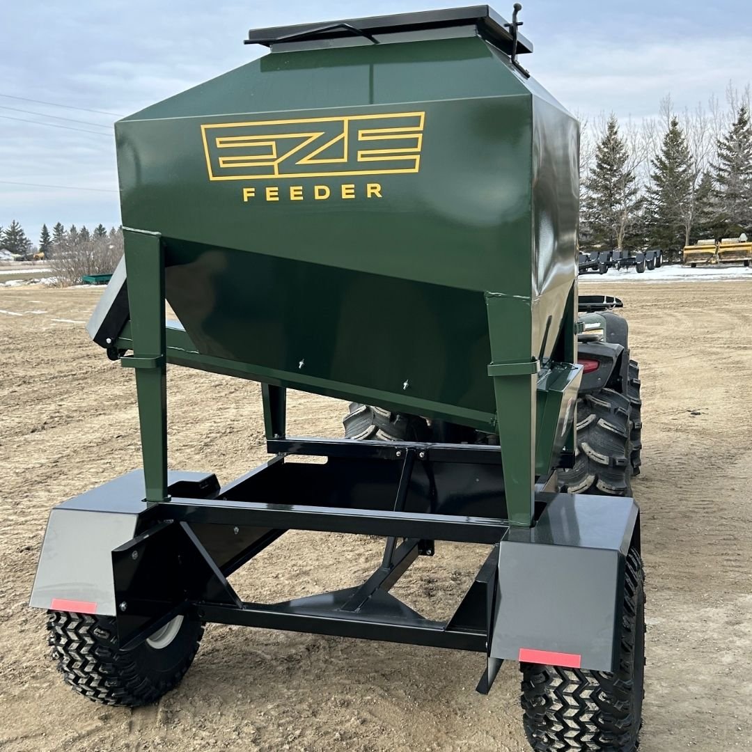 Green UTV15 tow behind Feeder hooked up to a grey quad / ATV  in an open field, trees in the foreground and a blue sky with some clouds.