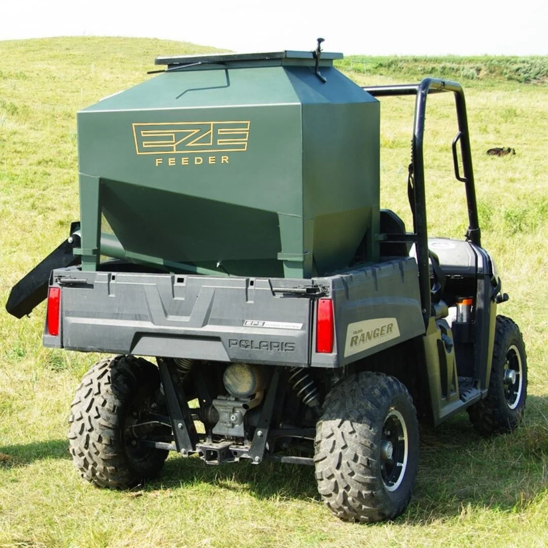 A green EZE Feeder portable feeder in the back of a UTV with the spout extended to discharge feed.