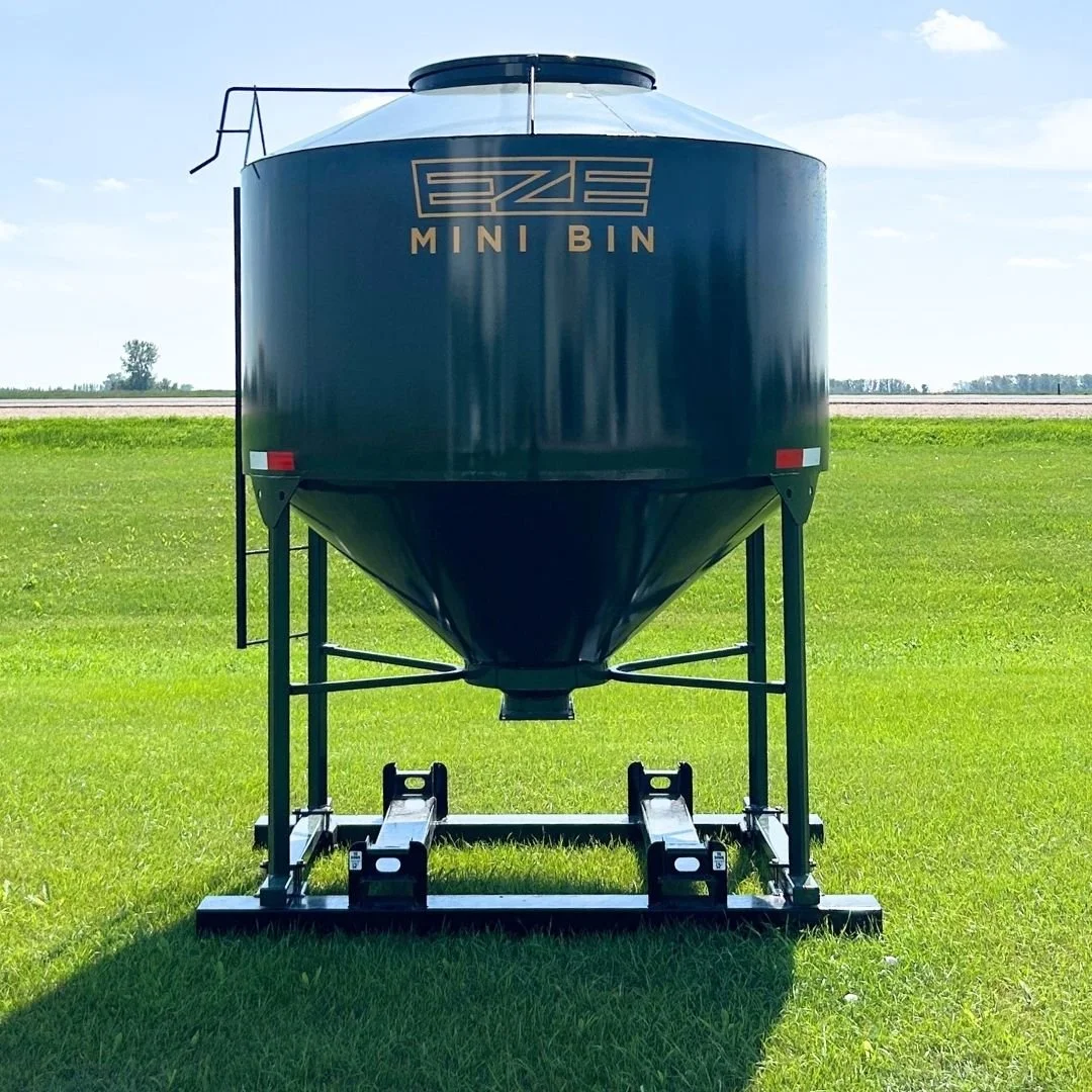 Front view of a 95 bushel feed storage bin, dark green with yellow logo, sitting on green grass with a blue sky.