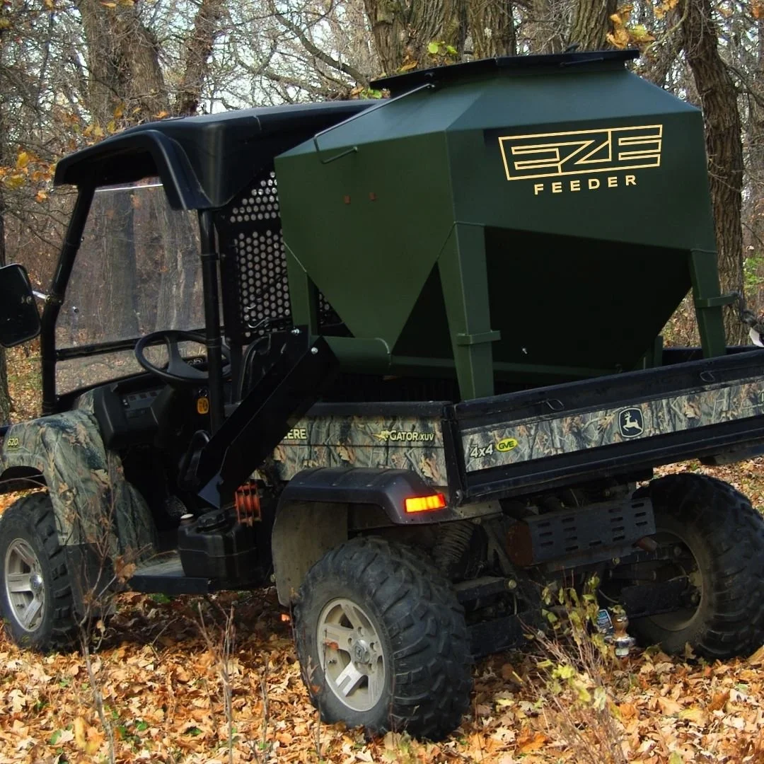 A utility terrain vehicle - UTV - with a green eze-feeder in the back located in a foresty area with autum leavs on the ground.