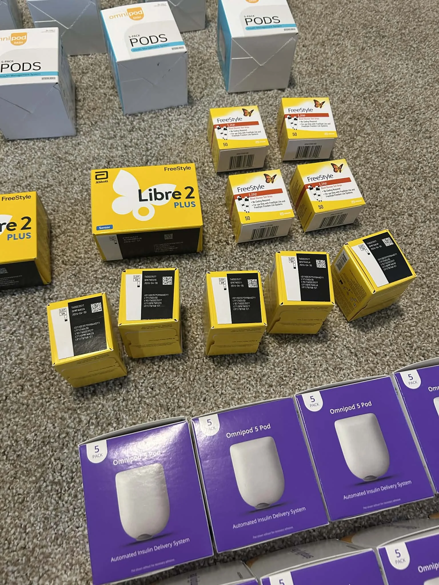 Multiple boxes of Libre sensors, Omnipod pods, and test strips laid out showing unused diabetic supplies