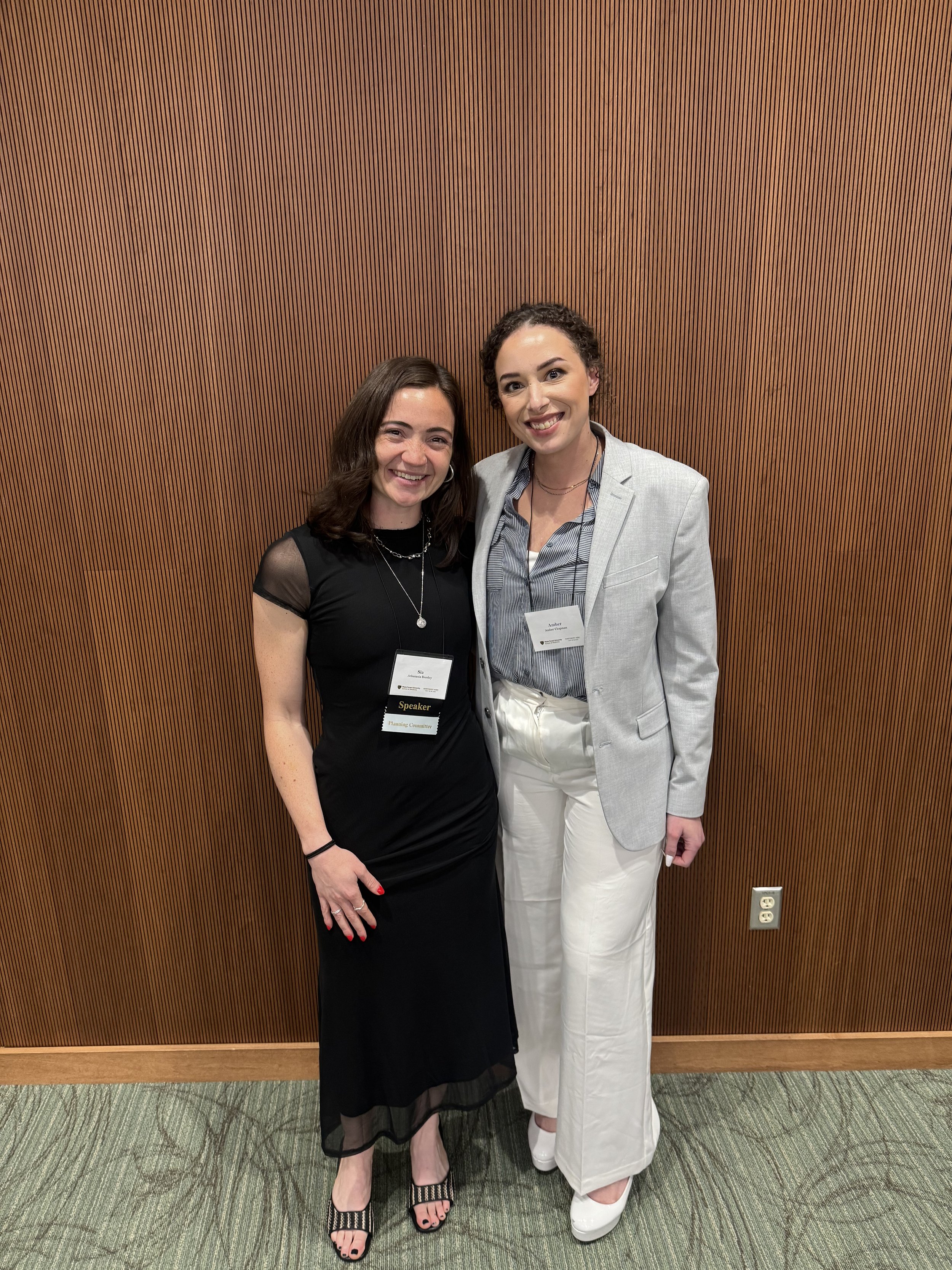 Two women standing together, smiling, in front of a wooden wall. One is wearing a black dress with sheer sleeves, black high-heeled sandals, and jewelry. The other is wearing a light gray blazer over a striped shirt, white pants, and white shoes. They are at a professional event, both wearing name tags.