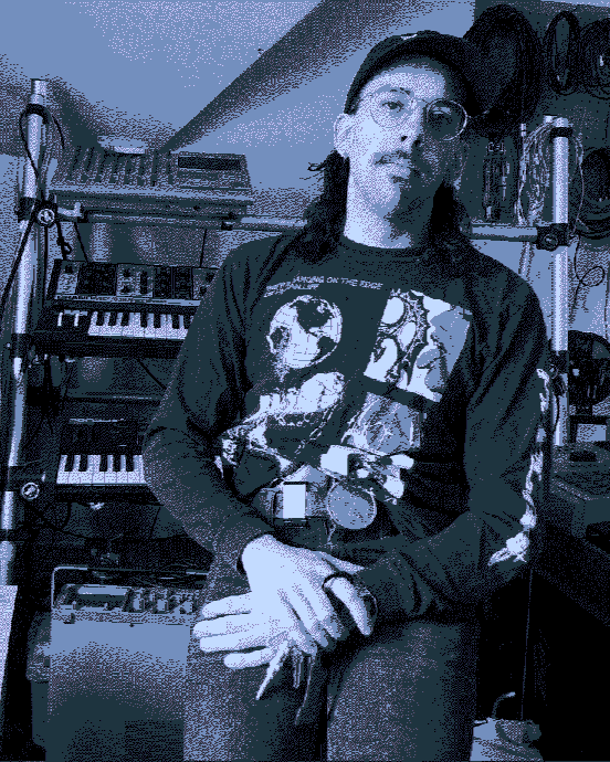 A person with long dark hair and glasses, wearing a graphic sweatshirt, standing in a music studio with keyboards, synthesizers, and music equipment in the background.