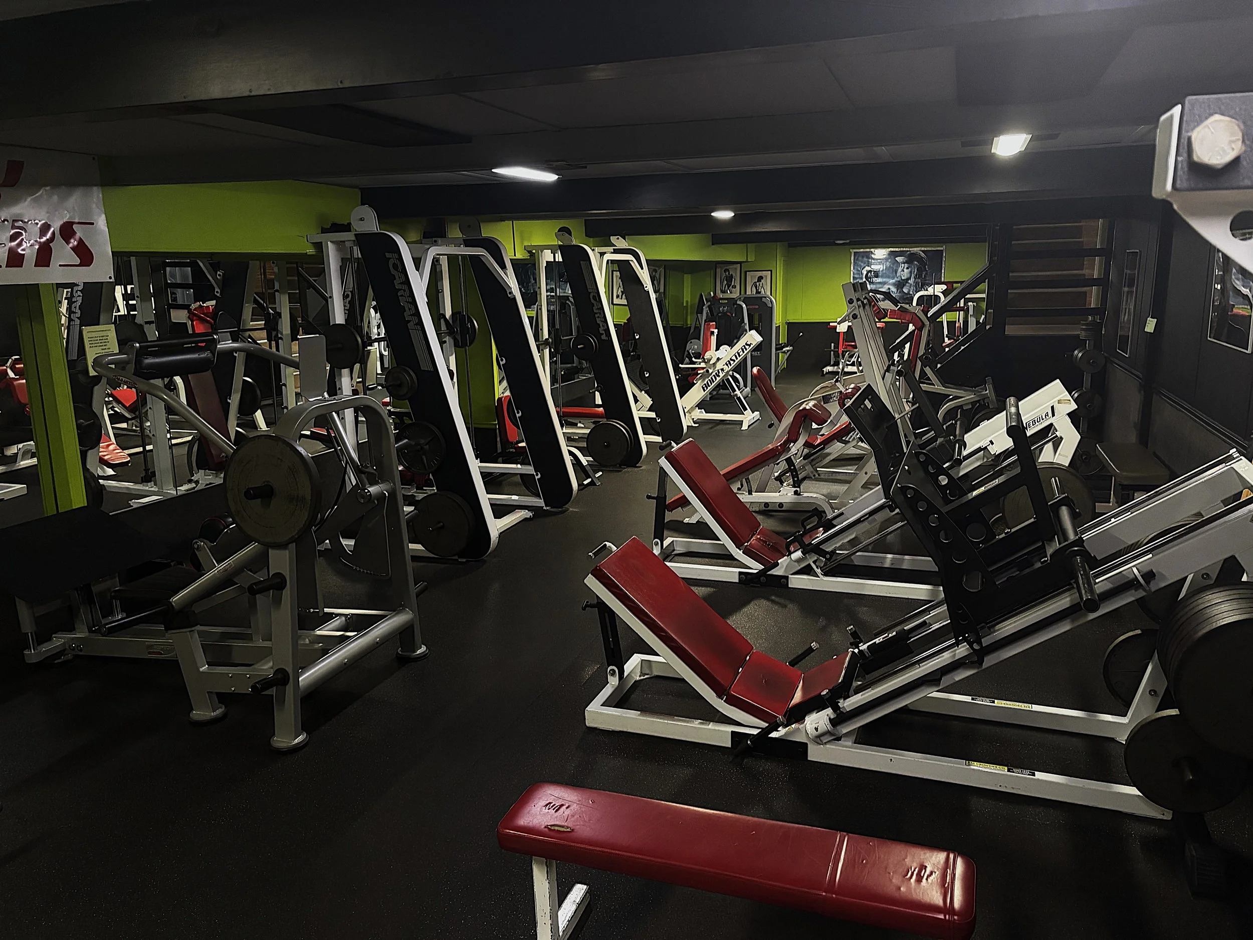 Interior of a gym with various weightlifting and exercise equipment, including benches, barbells, and machines, with black and green walls and ceiling lighting.