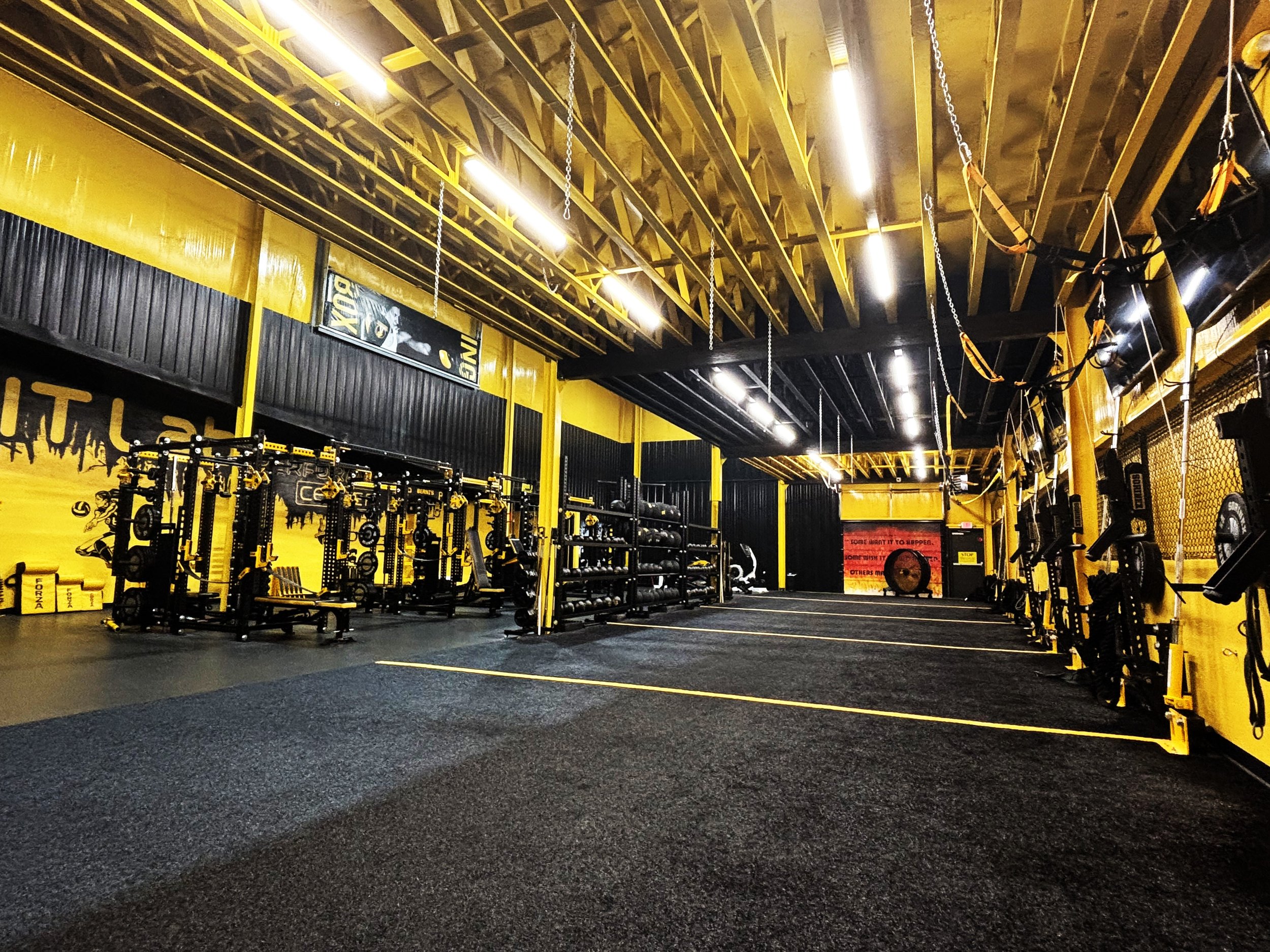 Interior of a gym with black and yellow workout equipment, including weight racks, benches, and TVs on the walls.