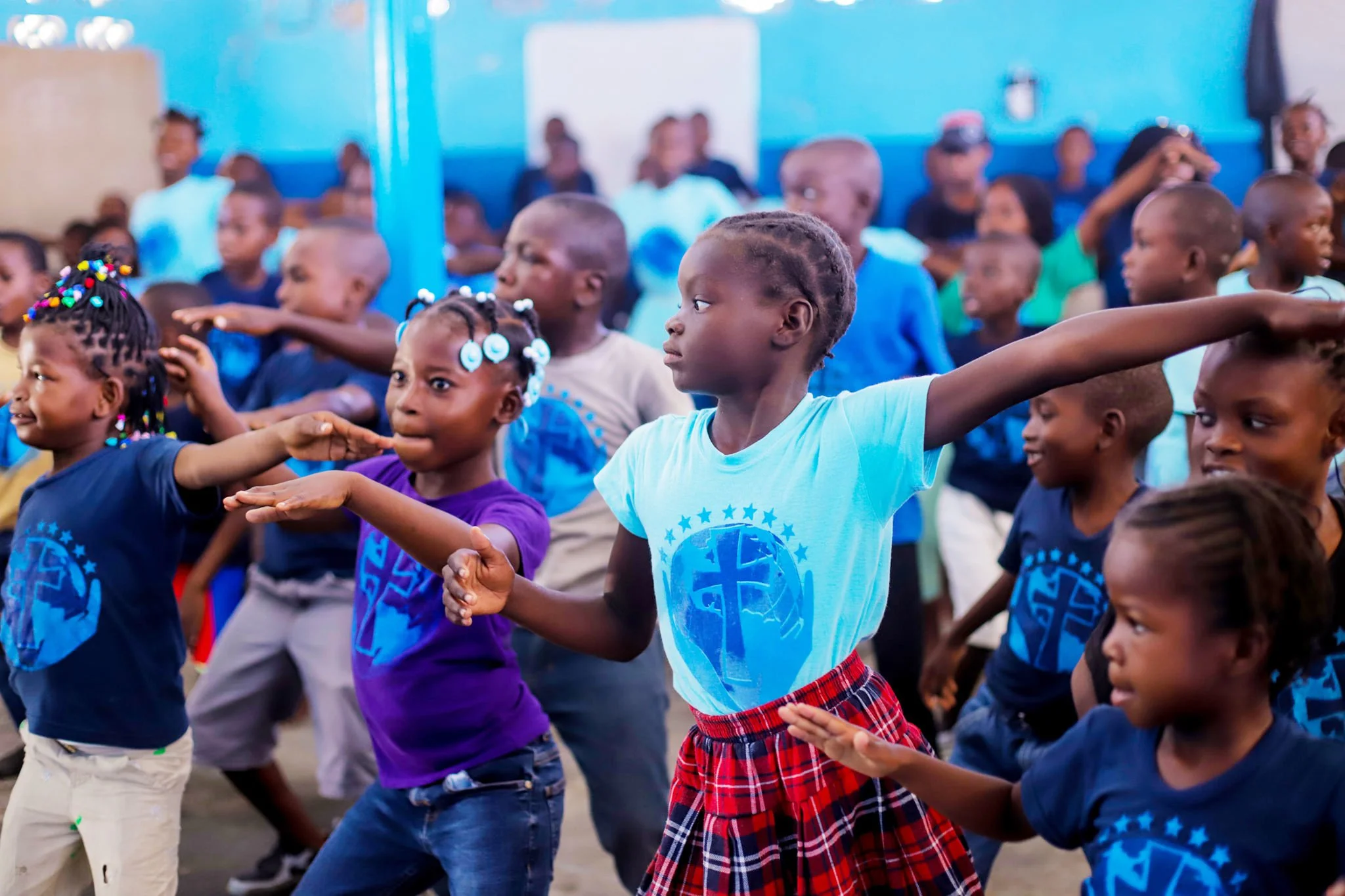 Children dancing together indoors, some with their arms extended or raised, wearing colorful clothes and smiling.