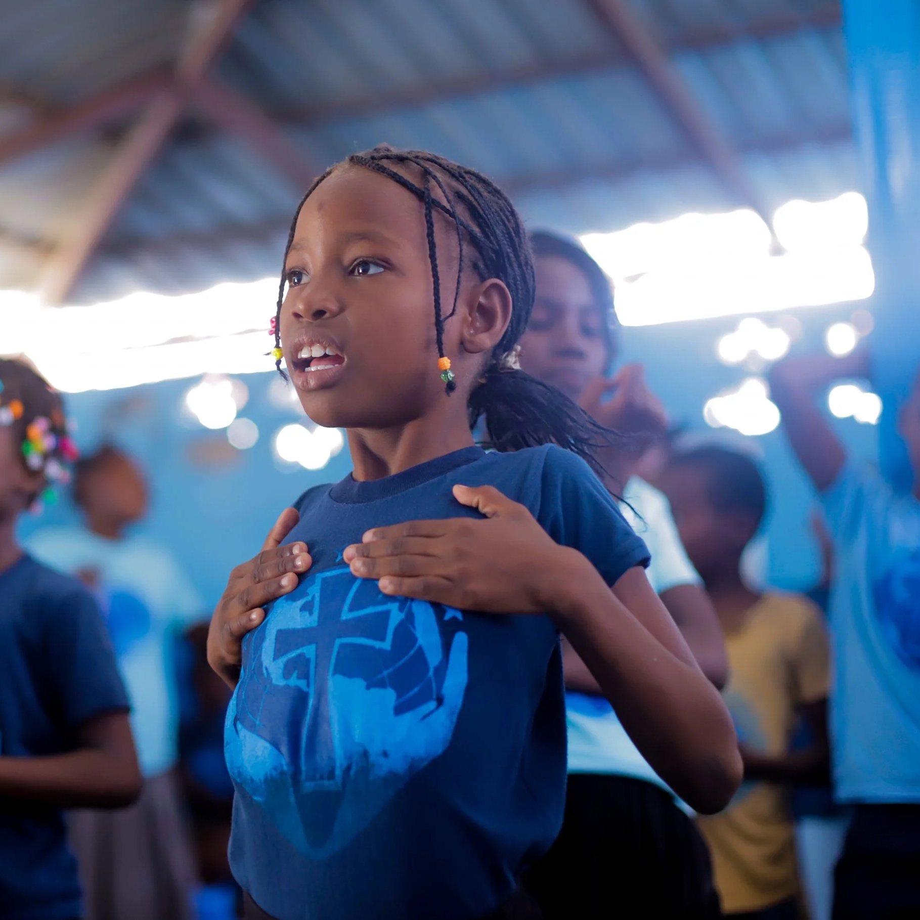 A young girl with braided hair and beaded accessories appears to be singing or reciting, with other children in the background inside a building with a thatched roof.