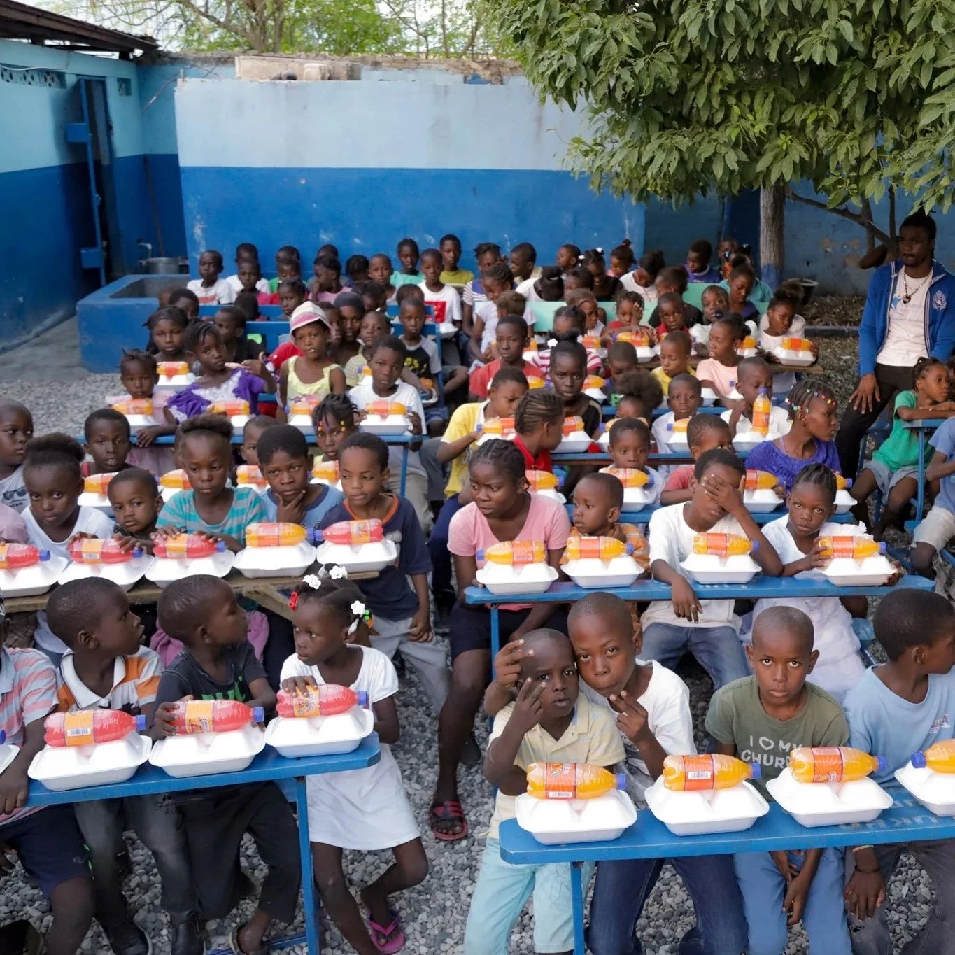 A large group of children sitting at desks outdoors, each with a meal consisting of a tray with food and a bottle of juice, in a courtyard area with blue walls and a tree.