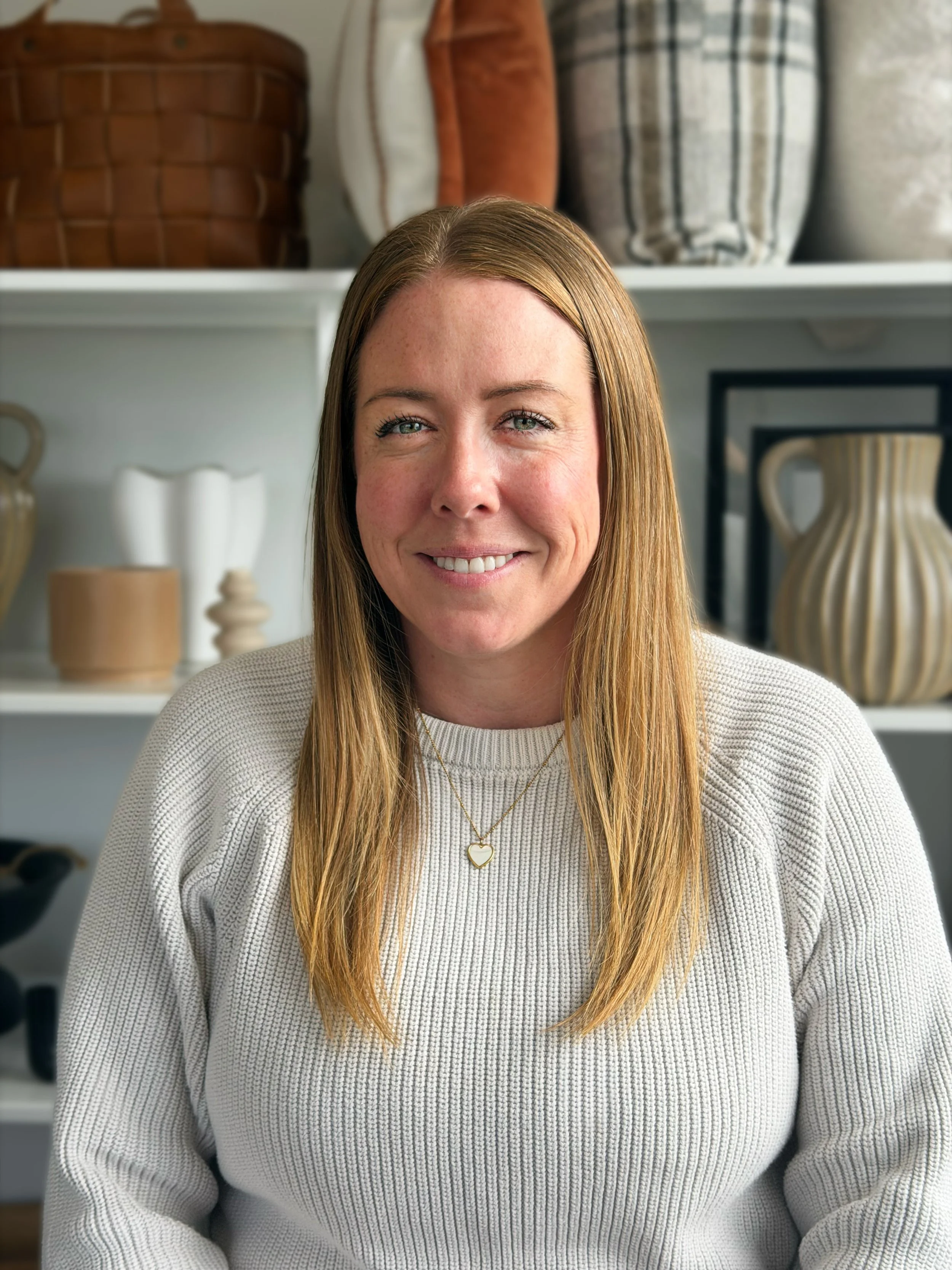 A woman with long, straight, red hair and blue eyes smiling at the camera. She is wearing a cream-colored sweater and a gold necklace with a heart-shaped pendant. The background shows shelves with decorative pottery and woven baskets.