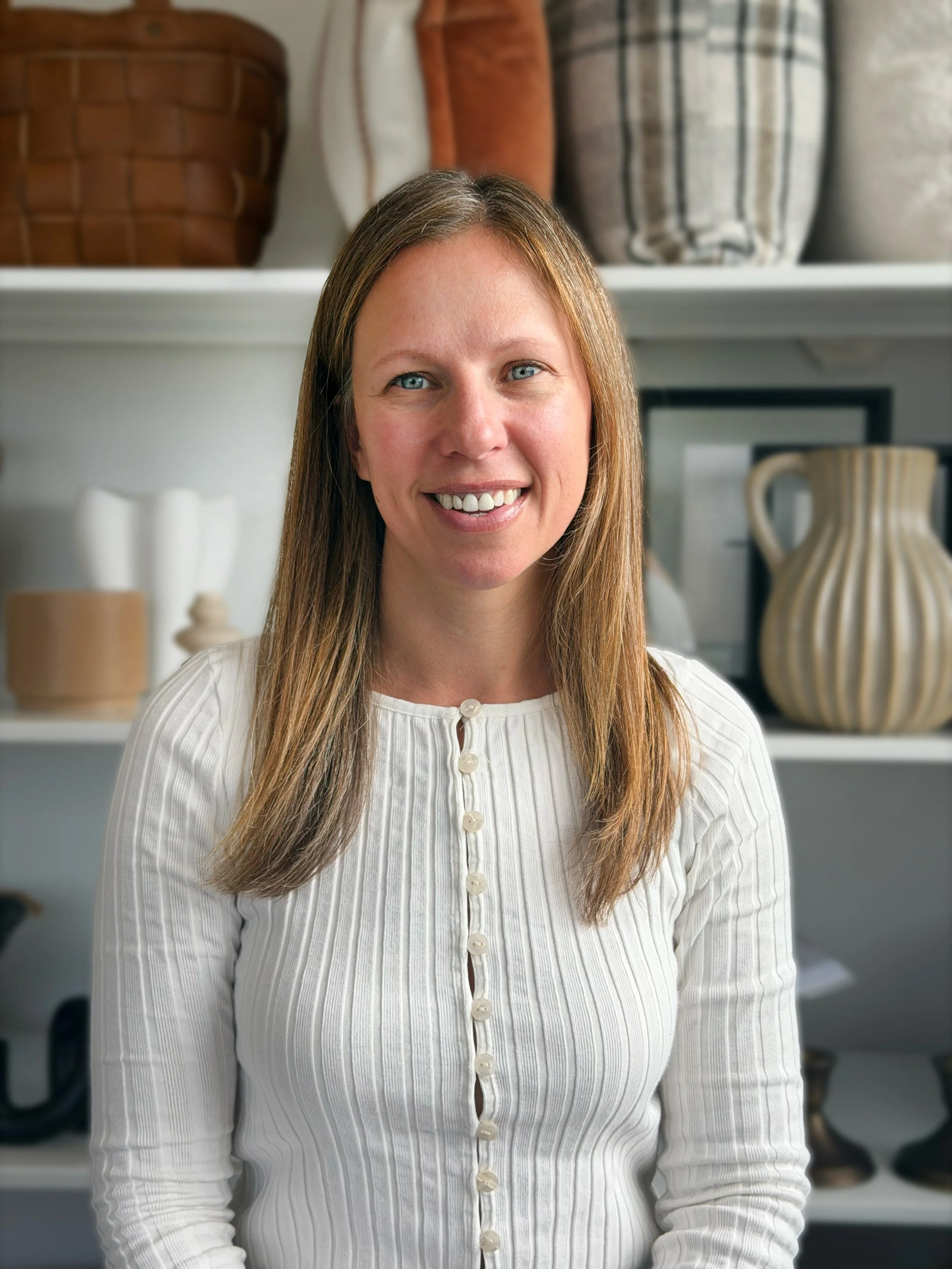 A woman with long light brown hair and blue eyes smiling, wearing a white button-up shirt with vertical stripes, standing in front of a white shelf with decorative vases and objects.