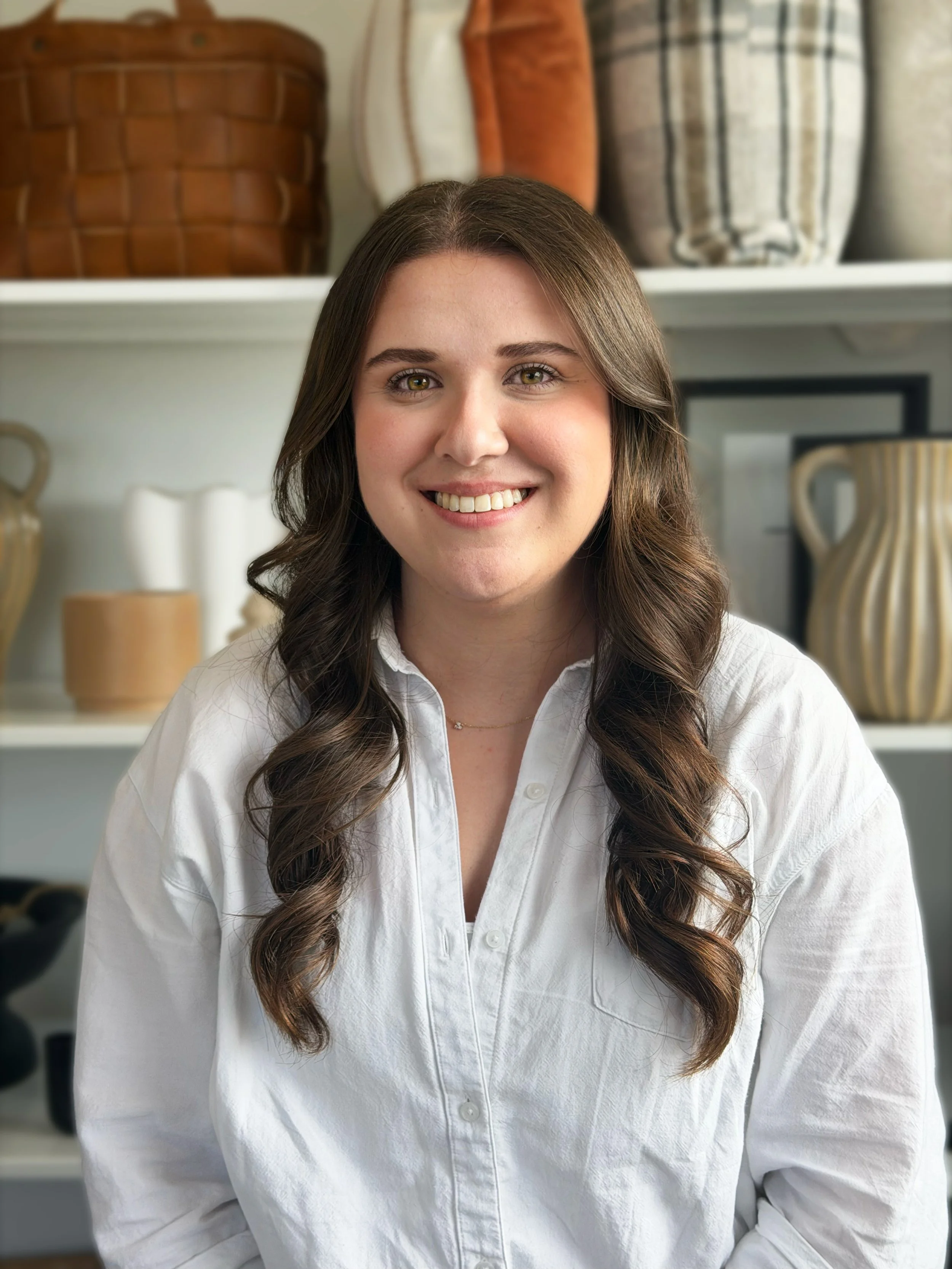 Young woman with long wavy brown hair, wearing a white button-up shirt, smiling, in front of a shelf with decorative pottery and plants.