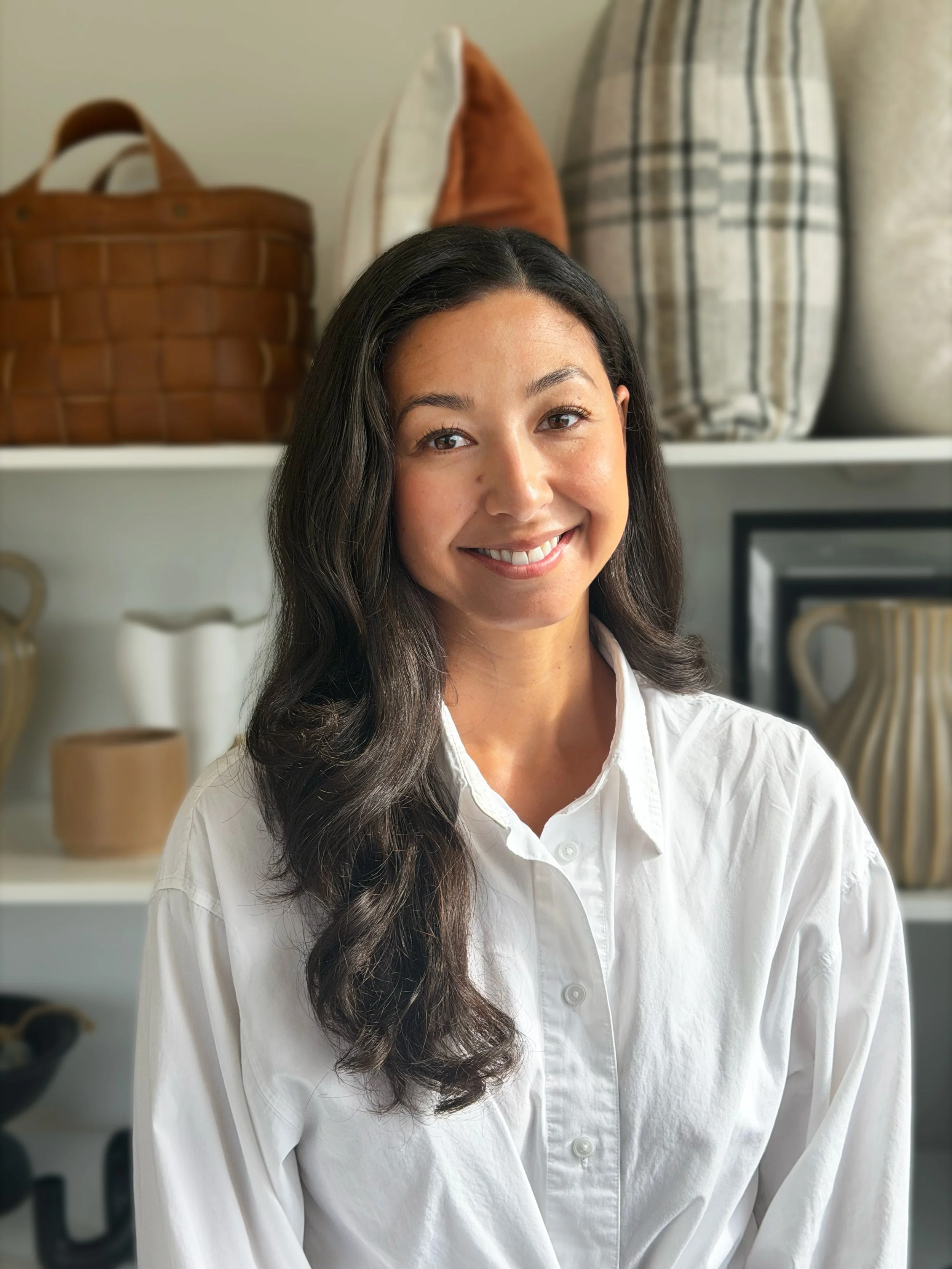 A woman with long dark hair smiling in front of a shelf with baskets and decorative items.