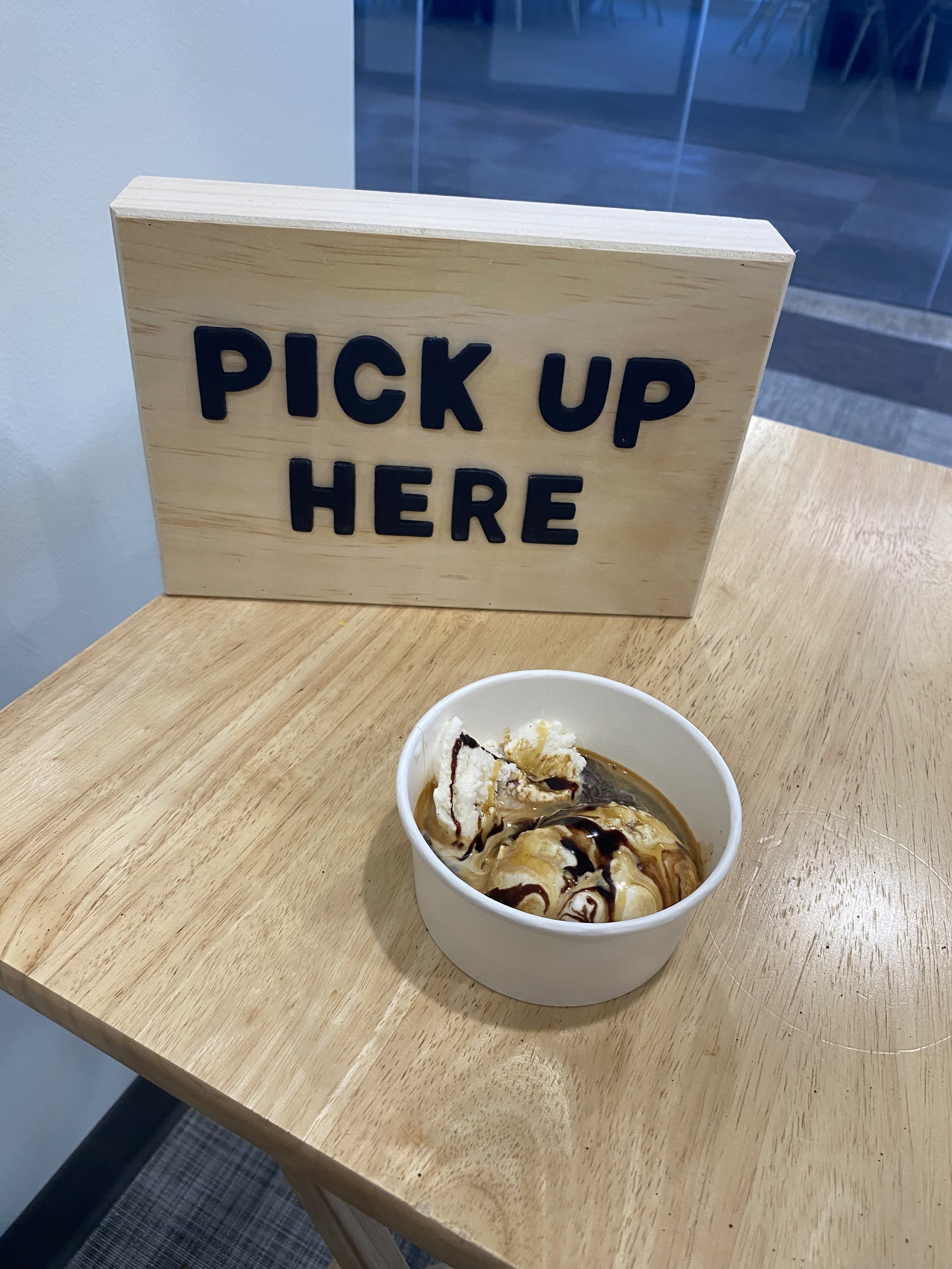 A small white bowl of ice cream with chocolate syrup on top and a piece of vanilla cake, on a wooden table next to a sign that says 'PICK UP HERE' in black letters.