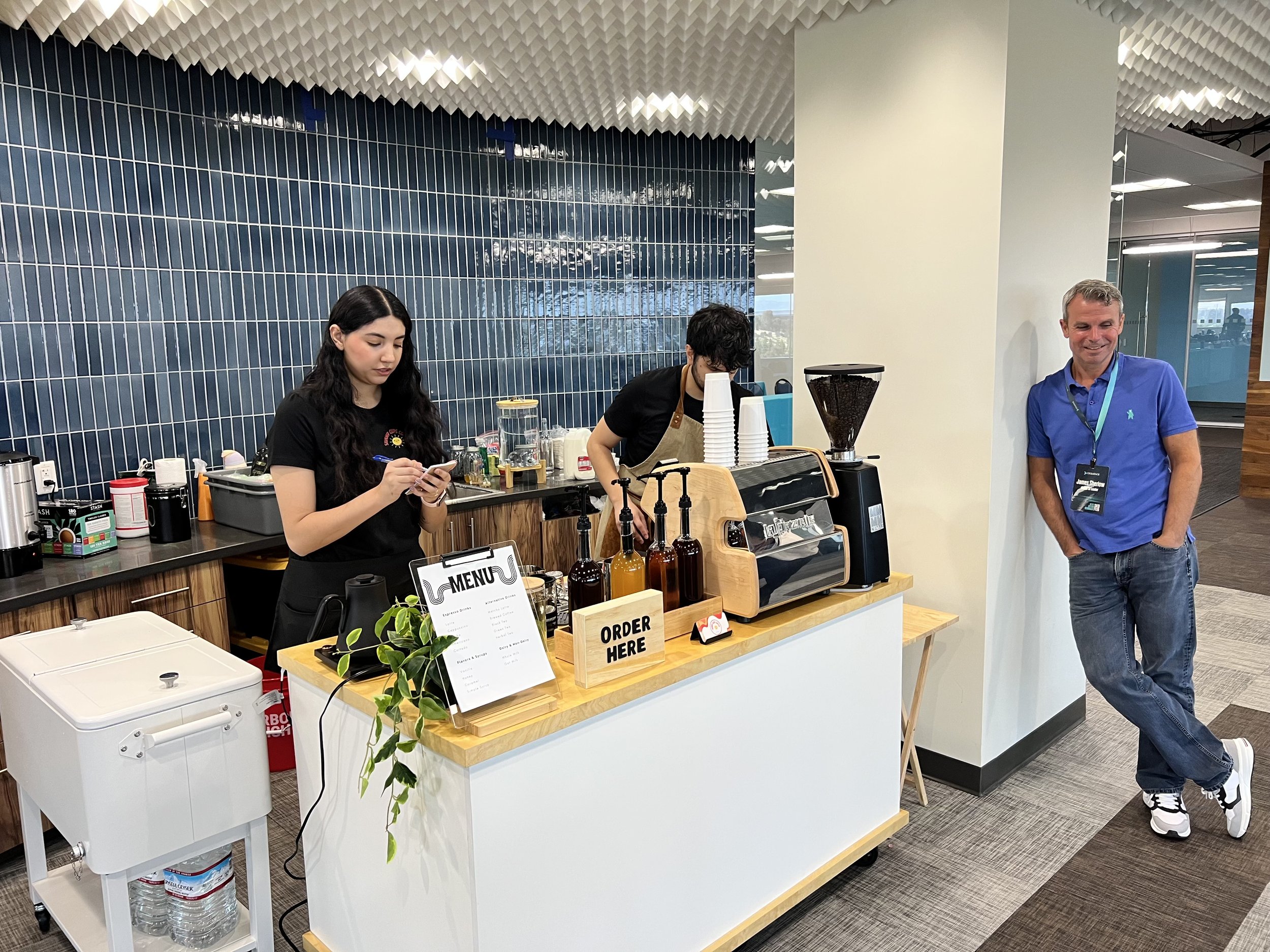 A coffee stand in an office or cafe, with two employees preparing drinks and a customer waiting. The stand has a menu, coffee equipment, bottles, and cups, with a man leaning against the wall smiling and chatting.