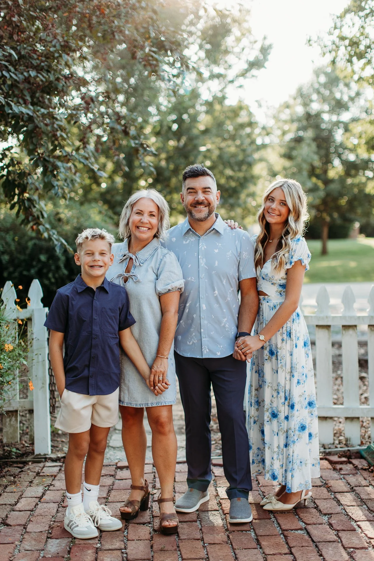 A multigenerational family portrait of five people outdoors, standing on brick pavement in front of a white picket fence with greenery and trees in the background, during daytime. They are smiling and holding hands.