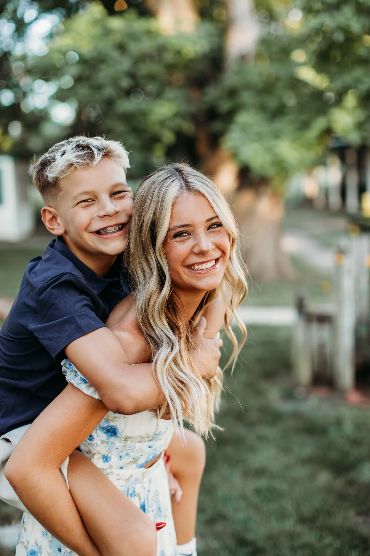 A smiling woman giving a piggyback ride to a young boy in a park, both looking happy and enjoying the outdoors.