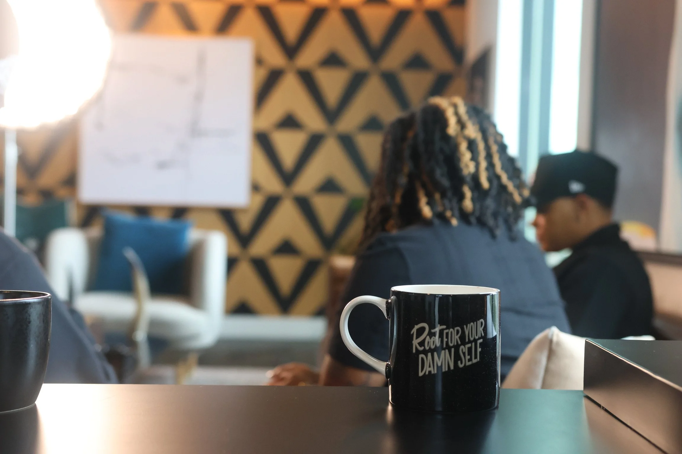 A black mug with the words 'Root for your damn self' on it is on a table in a cozy cafe. In the background, two people with curly and straight hair are sitting, facing away, with a patterned wall behind them and a large window letting in natural light.