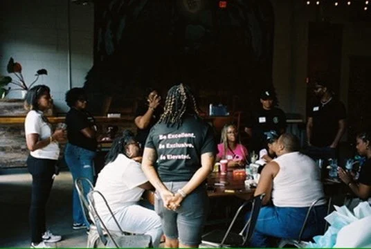 Group of people gathered around a table in a room, some standing and some sitting, engaged in a discussion or activity.