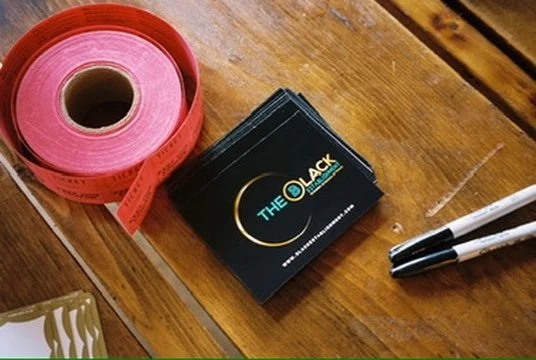 A rolled pink measuring tape, a small stack of black cards with gold and white text that reads 'The Black,' and two black and white markers on a wooden surface.