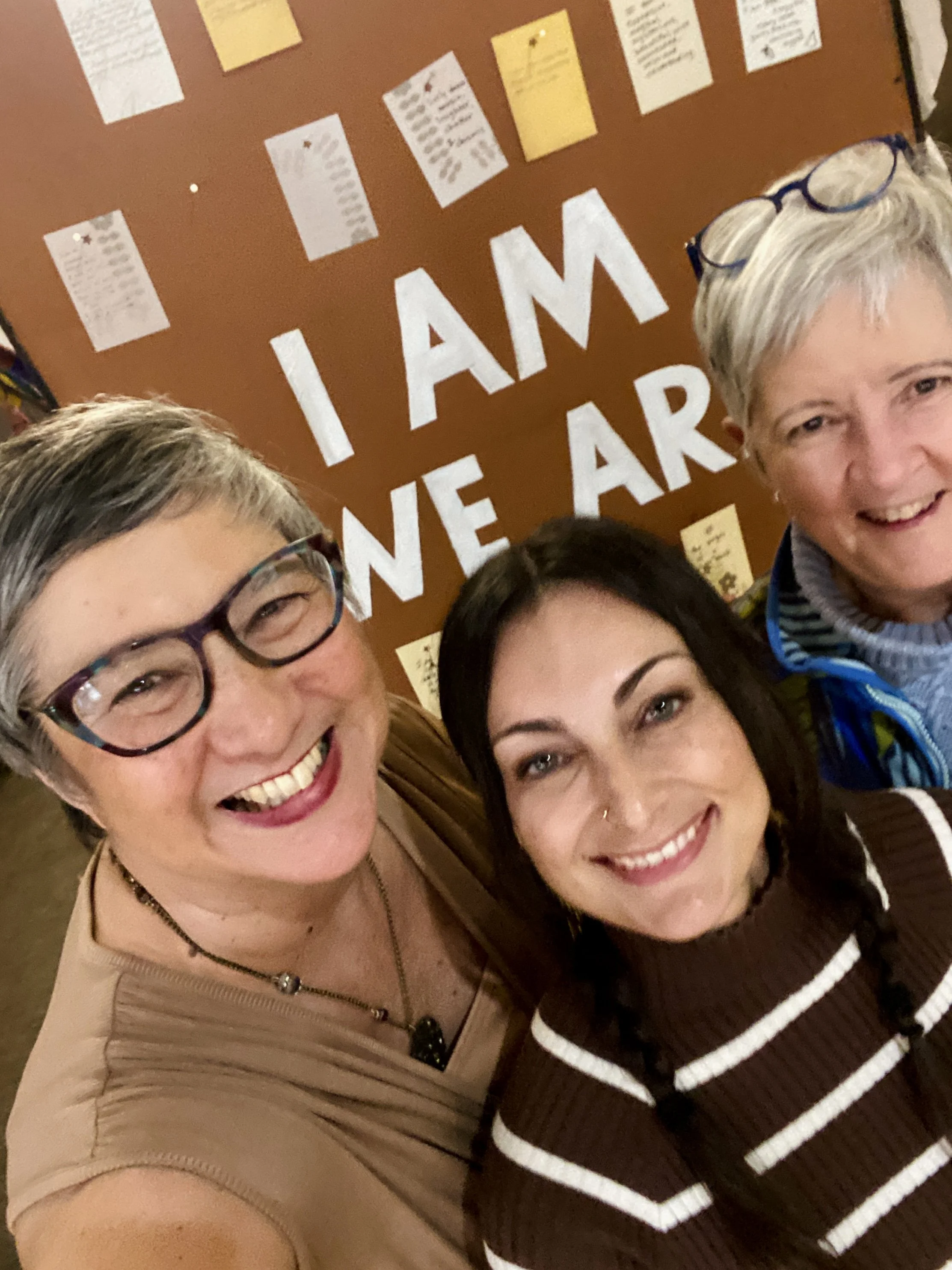 3 smiling women, retreat facilitators of the Rooted and Rested Retreat, standing  in front of a sign reading I Am/We Are