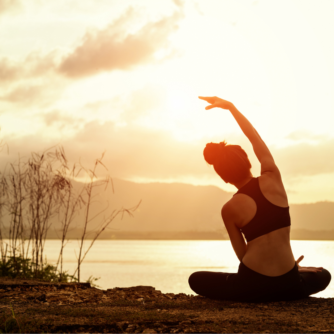 Woman in athletic clothing sits in a yoga pose while on an over look looking out over the water and mountains.