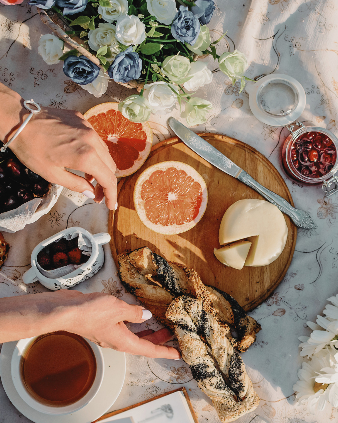 A table set for a picnic with a variety of foods including grapefruit halves, cheese, toasted bread with toppings, cherries, and a cup of tea, decorated with white and blue flowers.