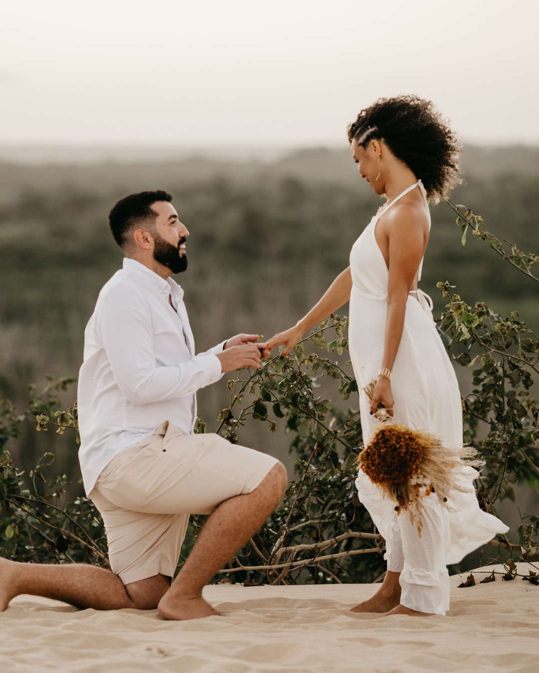 A man proposing marriage to a woman on a sandy beach during sunset, with the man on one knee holding a ring and the woman holding a bouquet, both smiling.