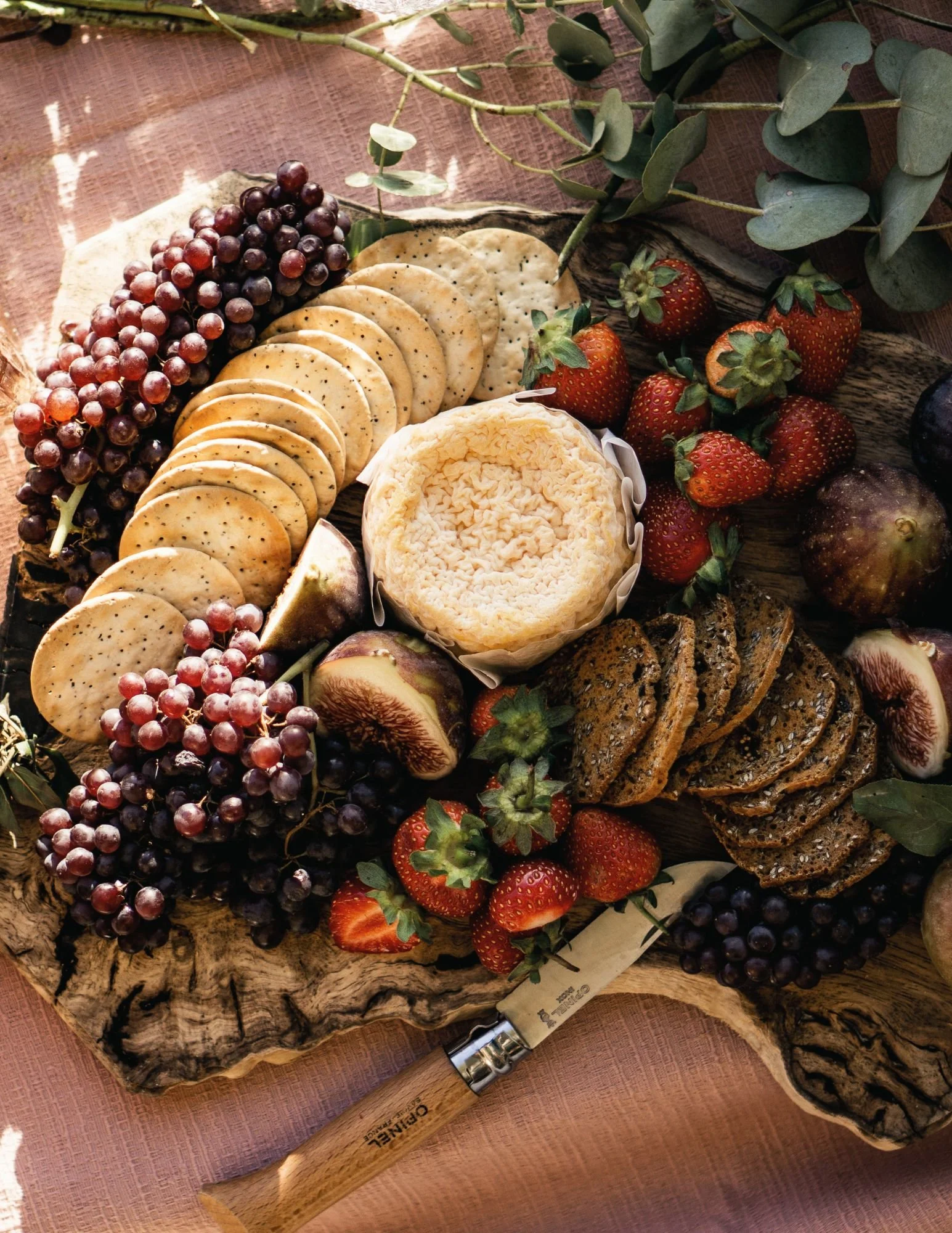 A wood platter with grapes, crackers, strawberries, a wheel of cheese, figs, and leafy sprigs, with a cheese knife and a wooden-handled kitchen spoon nearby.