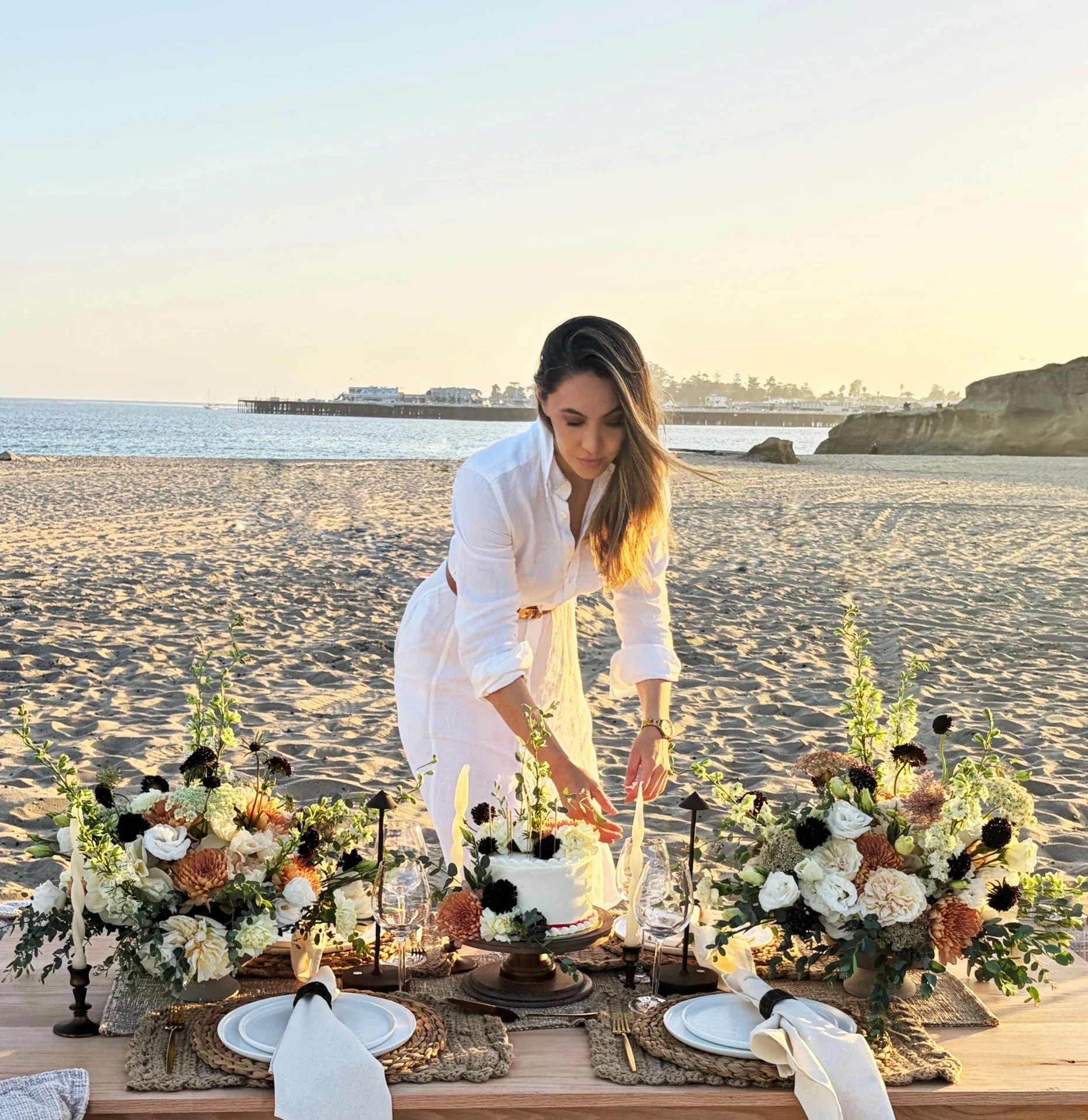 A woman owner setting up a luxury picnic on the beach at sunset. With elaborate table settings and extravagant floral arrangements.