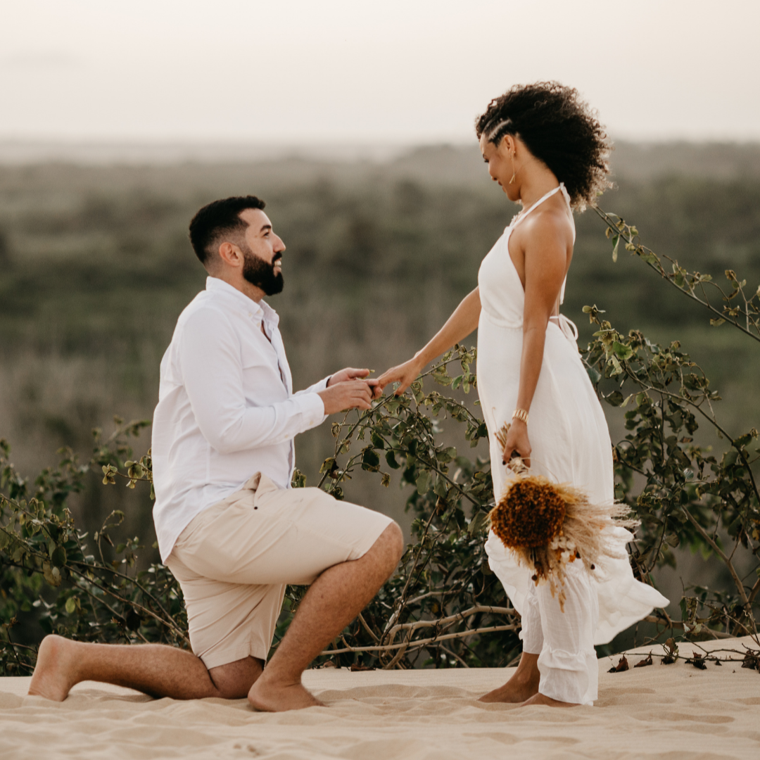 Man with a dark beard proposes while down on one knee. Woman in white dress holds mans hand and holds flower boquet in the other. The couple stands on the sand with rolling greenery behind them.