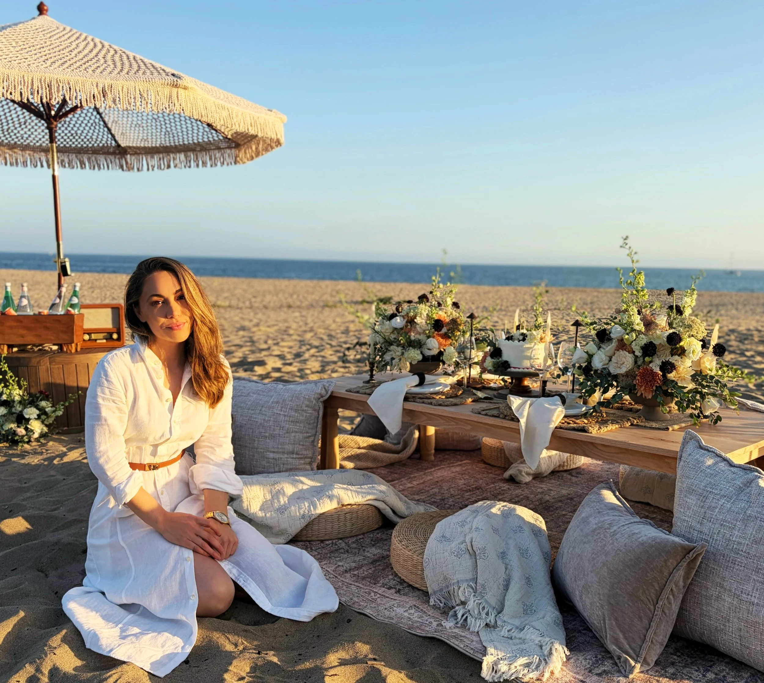Woman owner sitting in front of picnic on the beach with cake, refreshments, an umbrella, and an old music speaker. The ocean is in the background. There are rugs, pillows and blankets around the table, creating a cozy atmosphere.