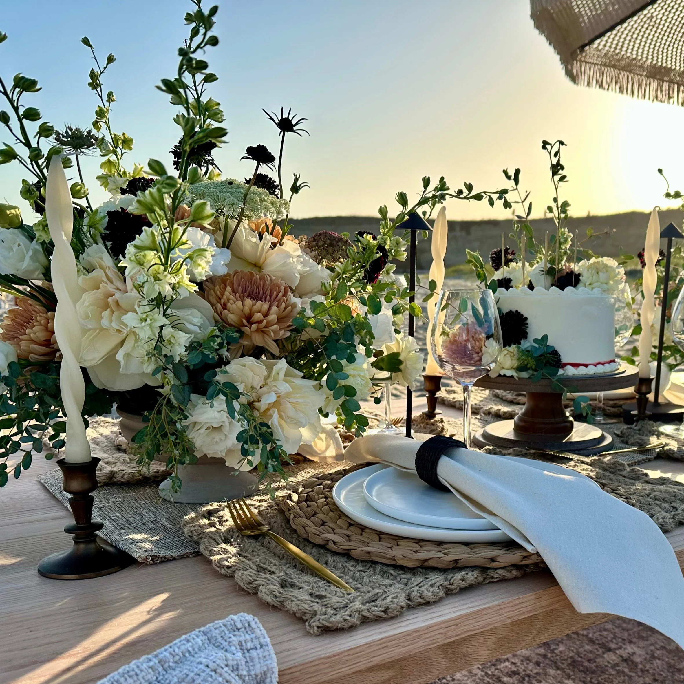 Picnic table with elaborate floral arrangements. place settings and a white cake with flowers on it. Sunset on the beach.