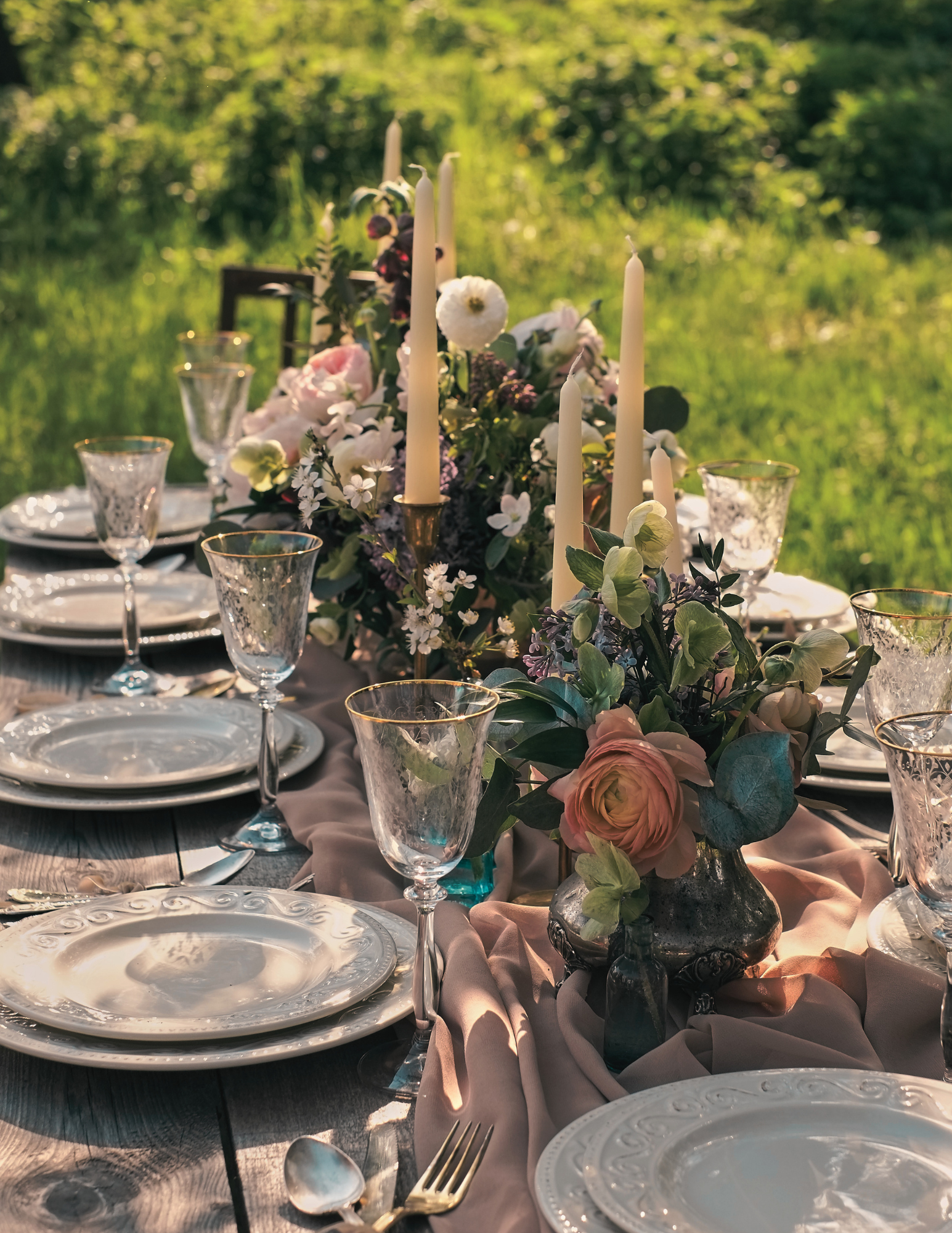 Outdoor table setting, with elegantly styled florals. Natural candles with brass candlestick holders, and neutral soft tableware. Dappled light shines down through trees in a back yard.