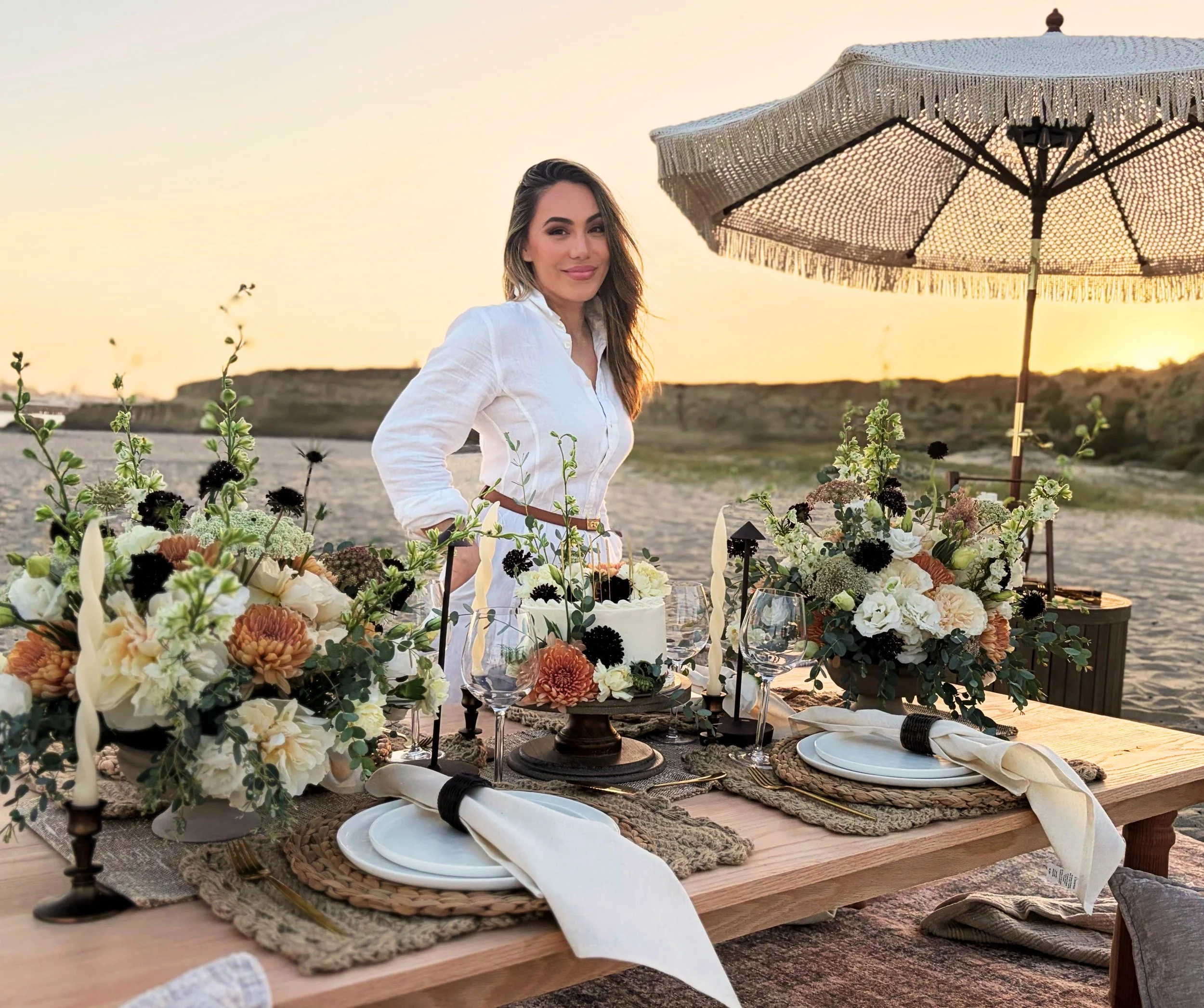 A woman owner standing on a beach in front of a luxury picnic table with place settings, a cake and elaborate flower arrangements. An umbrella sits in the background with a warm sunset.