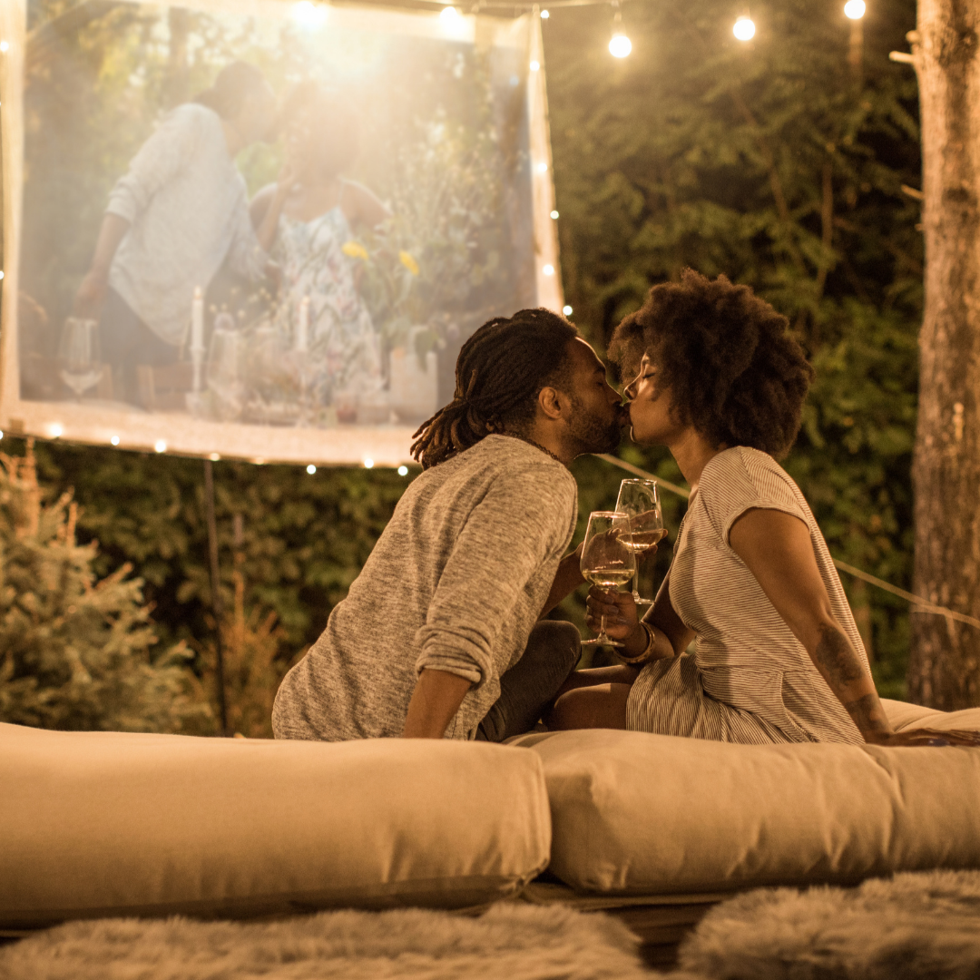 A couple sits on lush pillows and blankets in the back yard at night. In the background there is a back yard movie screen showing memories. The couple shares a kiss while enjoying white wine.String lights hang above creating a romantic glow.