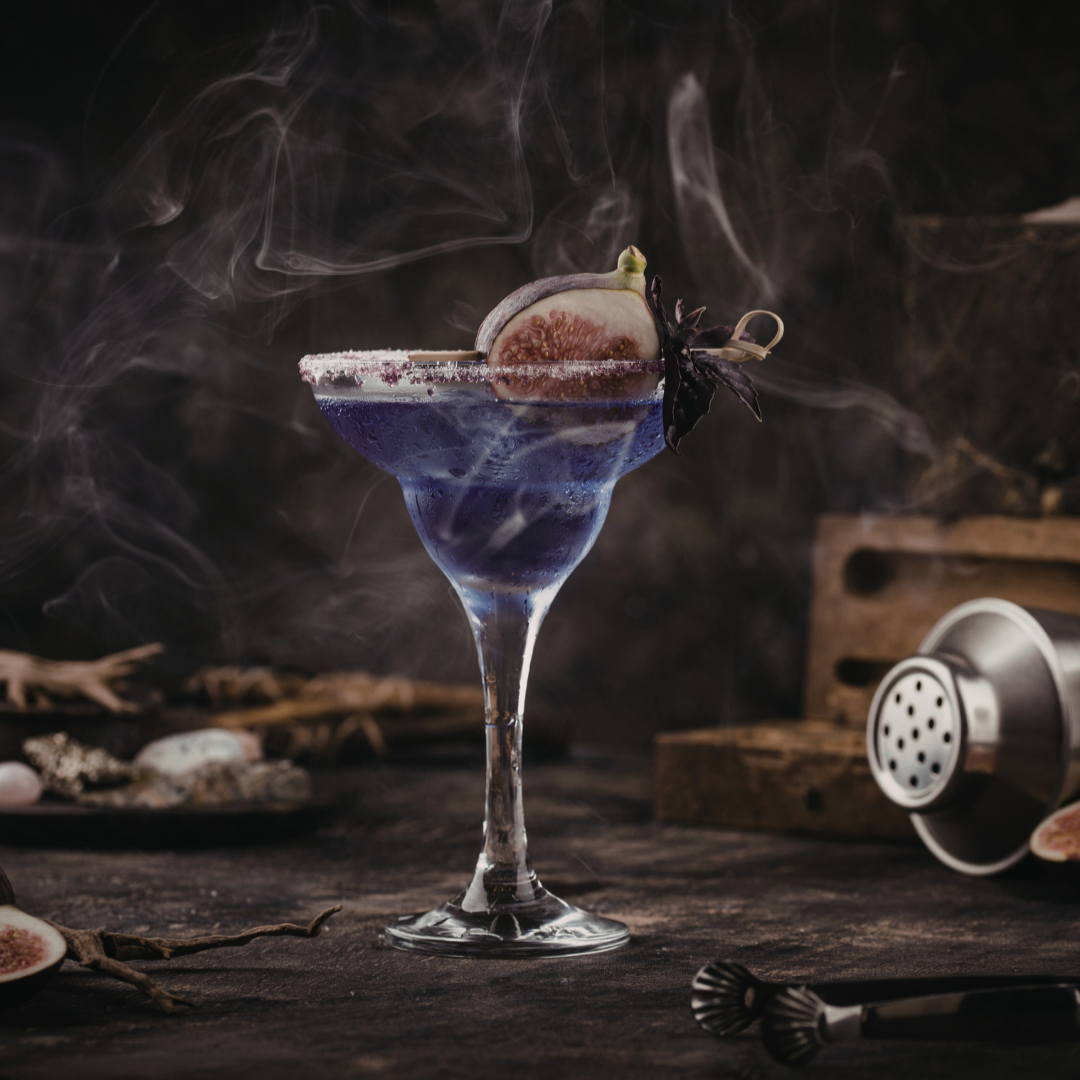 Dark picture of a sugar rimmed cocktail glass with a purple colored drink inside. A fig sits on top of the drink as a garnish. Smoke dances around the picture.