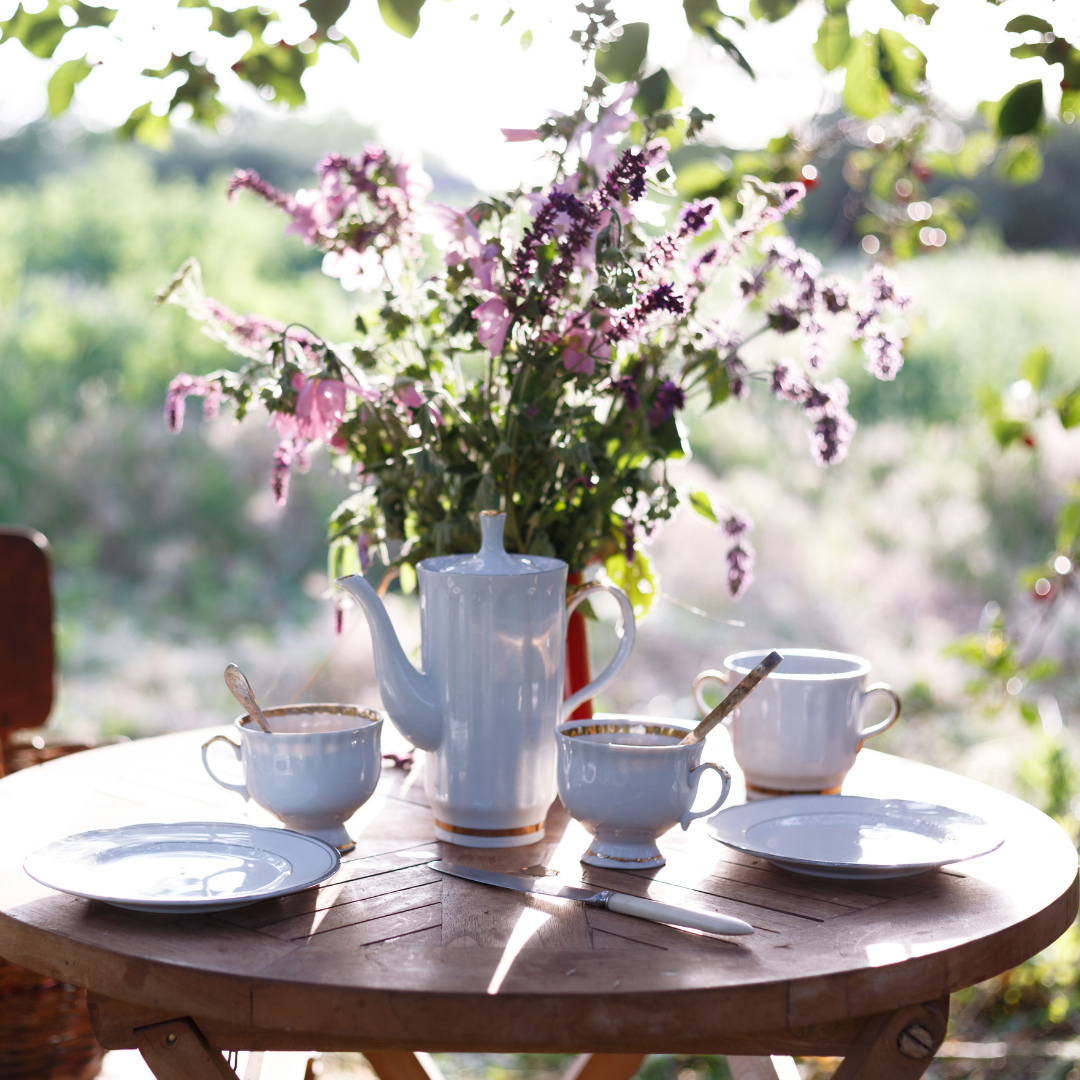 Outdoor table with white tea setting. Flower vase holds purple flowers. Dappled sun shines through trees. The background is a sunny field.